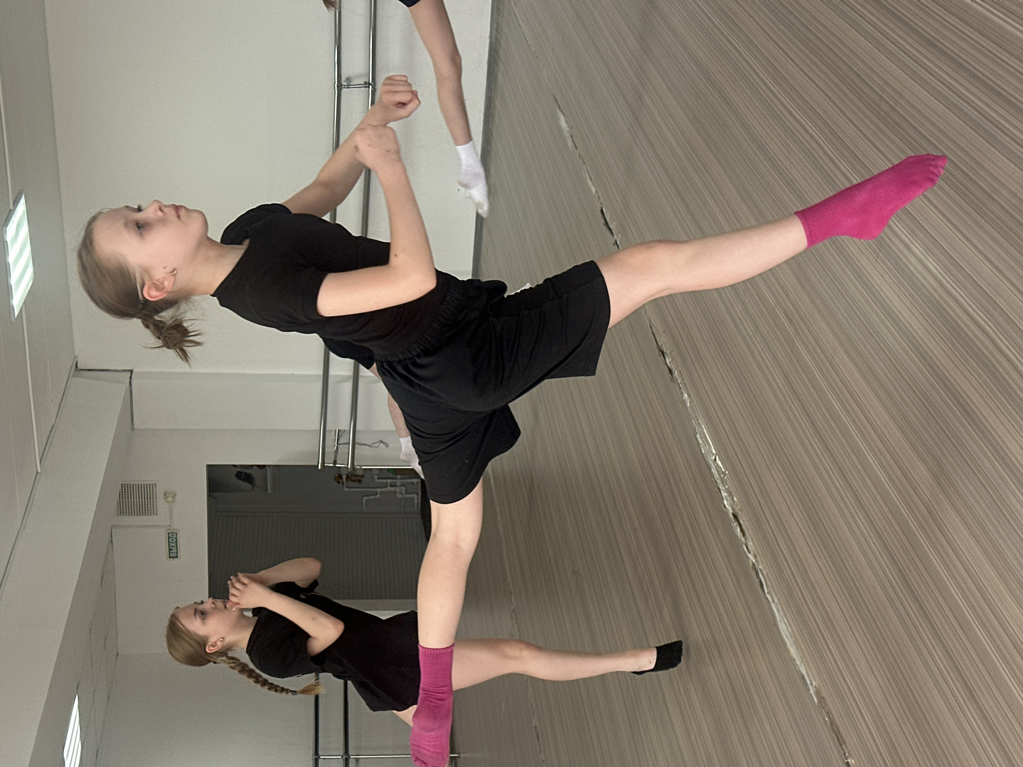 The image depicts two young girls in a dance studio setting, likely in Votkinsk, Russia. The foreground shows a girl in black shorts and a t-shirt, wearing bright pink socks and extending one leg. Behind her, a similar scene is mirrored with another girl in identical attire, similarly positioned. Additional figures are partially visible in the background, suggesting a group activity. The studio has mirrors, a barre, and what appears to be wood-grain flooring. A door with an exit sign is visible.
