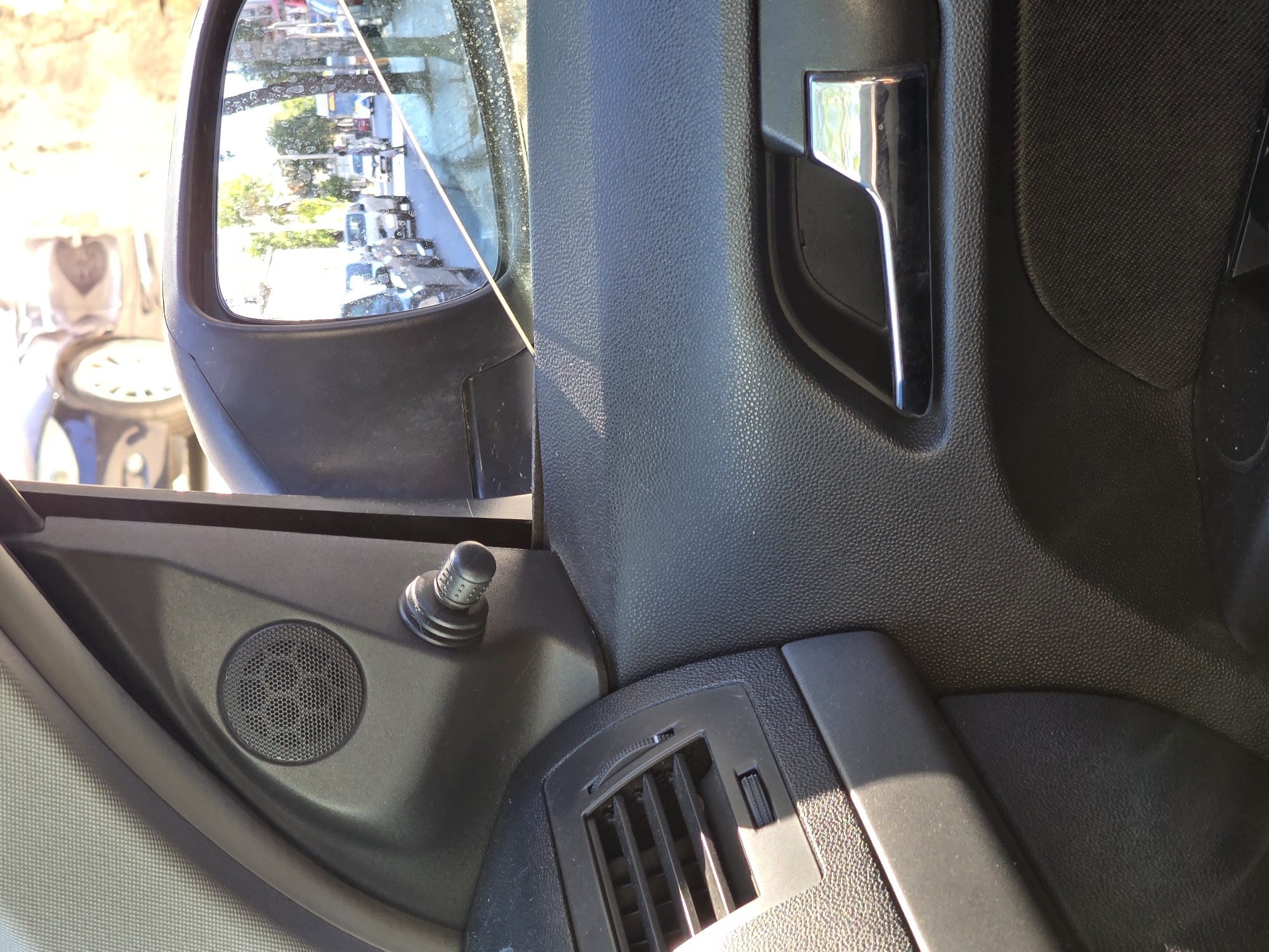 The image captures a close-up view of the interior of a car, focusing on the driver's side door panel and a side-view mirror. The door panel is made of black textured plastic, with a prominent chrome door handle and a speaker grill near the window. To the left of the speaker is an air conditioning vent. The side-view mirror reflects a street scene with several cars in traffic, trees lining the road, and a bus in the distance. Sunlight streams into the car, casting shadows on the interior surfaces. The context of Athens, Greece, is not evident from the visual information provided within the image itself. There are no people visible within the frame of the car's interior or clearly identifiable in the reflection.