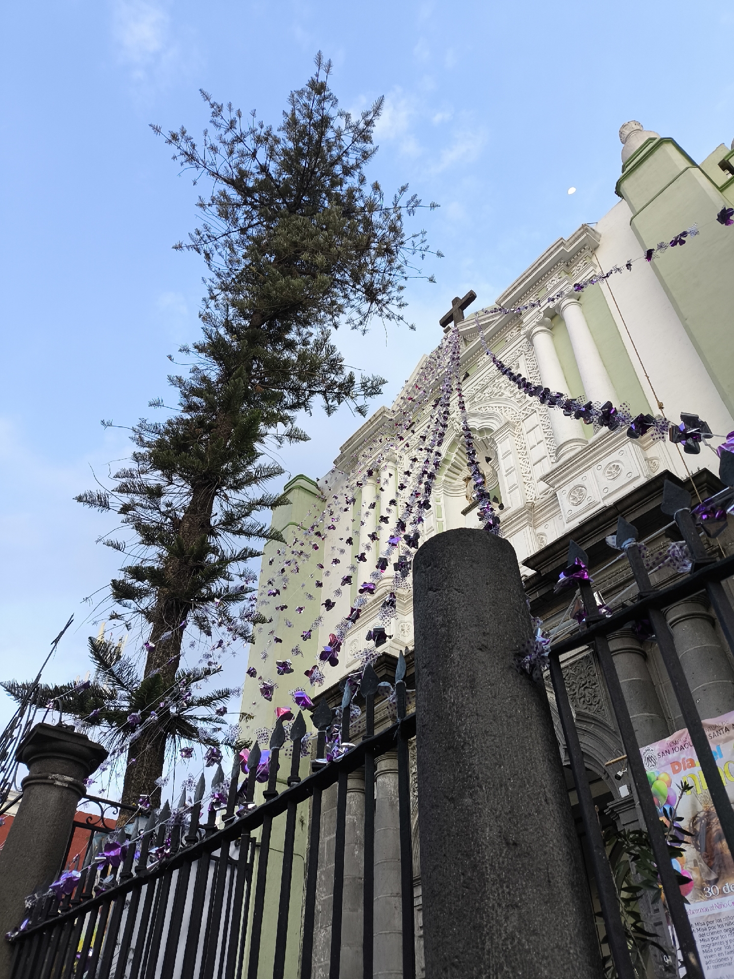 The image captures an upward-angled view of the Templo de San Joaquín, a church in Puebla, Mexico. The building has a white facade with columns, arches, and ornamental details. The church is decorated with strands of purple ribbons or flowers. A tall pine tree grows to the left of the church, and its branches are intertwined with the same purple decorations. A dark metal fence runs in front of the church, with the same purple decorations. A stone post is positioned prominently in the foreground. A small segment of the sky is visible, with a very small moon. A poster is attached to the side of the church.
