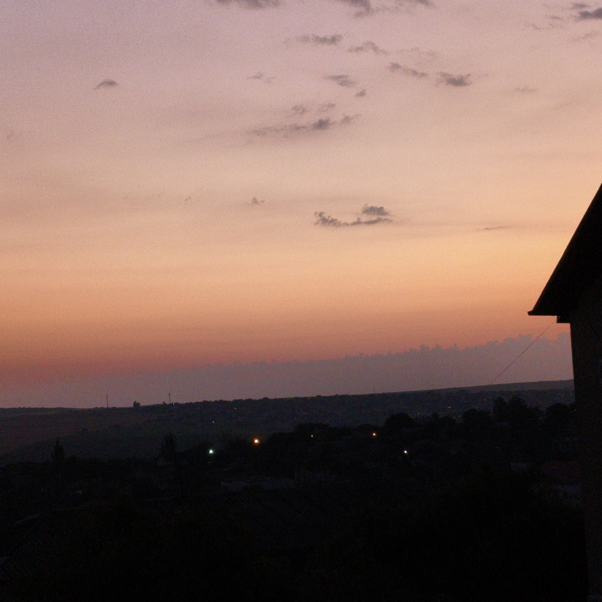 The image depicts a silhouetted landscape at dusk or dawn. The sky is a gradient of soft pinks and oranges, with a few scattered, wispy clouds. In the foreground, the dark silhouette of a building's roofline is visible on the right side of the frame. Beyond this, a dark, undulating horizon line suggests hills or a varied terrain. Small, faint lights are visible in the distance, dotting the dark landscape, indicating the presence of a settlement or populated area. The overall impression is one of quiet and transition between day and night.