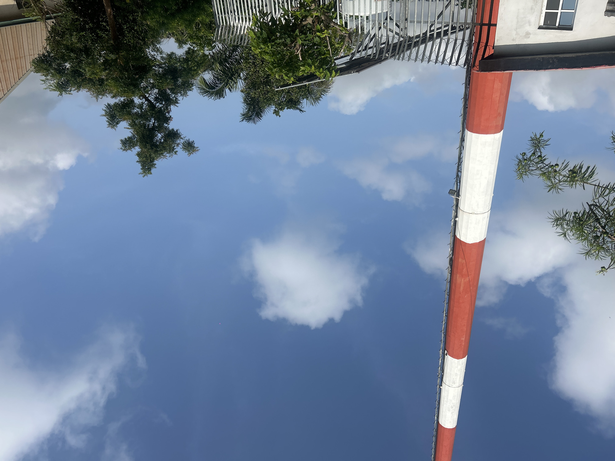 In Masaka, Nigeria, a tall, red and white striped tower, possibly a radio or communication tower, stands against a bright blue sky with scattered white clouds. A section of a light-colored building with a dark-pitched roof and a window is visible in the lower left corner, partially obscured by the tower. A metal fence with barbed wire runs horizontally across the middle ground. Beyond the fence, lush green trees and palm trees create a dense foliage backdrop. The scene suggests an outdoor industrial or infrastructural setting within the city.