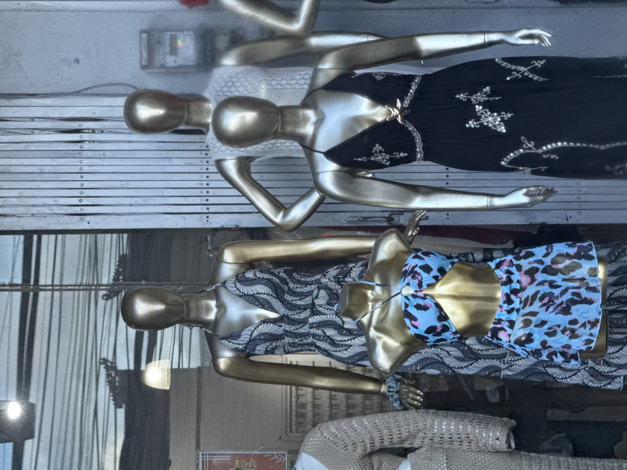 The image captures a retail shop window display featuring four golden, headless mannequins, arranged in two rows. The image is oriented vertically, showing the mannequins sideways.
The two mannequins in the upper part of the image display different outfits. One wears a white, intricately lacy, short-sleeved top. Next to it, a mannequin is dressed in a black spaghetti-strap top or dress adorned with elaborate silver embroidery, featuring star-like and swirling patterns.
The two mannequins in the lower section showcase distinct apparel. One is clad in a dark patterned dress, possibly a jumpsuit, characterized by a wavy, feather-like design in shades of grey and white. The adjacent mannequin sports a vibrant light blue bikini top with a black and pink leopard print, complemented by a matching sheer sarong or cover-up. A light blue scrunchie is visible on its wrist.
The mannequins themselves are made of a reflective, golden material. Behind them, the background appears to be a light-colored, possibly white or grey, slatted structure, which could be an interior wall, a display panel, or a security shutter. In the upper background, a control panel with a small red indicator light is visible.
The lower portion of the image reveals significant reflections on the shop window glass, depicting the external environment. These reflections include vertical elements suggesting buildings or poles, along with trees and bright light sources, indicating the photograph was likely taken during daylight or early evening hours in an outdoor setting like North Kuta, Indonesia. The very bottom of the image shows folded or stacked knitted textiles, part of the shop's merchandise. No clear text is legible on any signs within the frame.