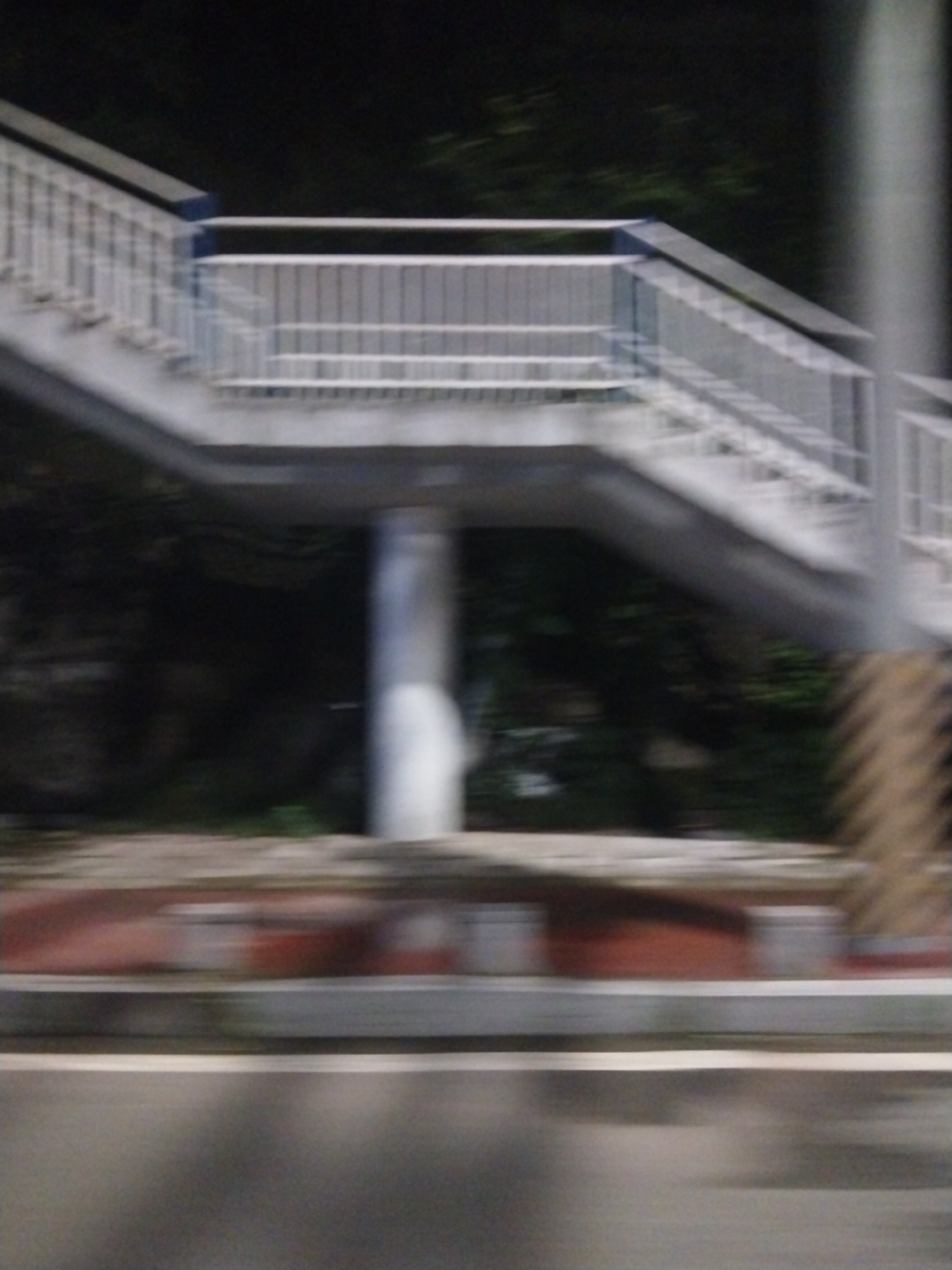 The image appears to be taken at night in what might be the Jiulongpo District, China, though this is speculative. There is a pedestrian overpass spanning a road. The overpass is constructed of metal with a white railing. It is supported by a central white pillar. The road is asphalt with a white line marking a lane. The image is blurry suggesting motion or poor focus. Vegetation is present behind the overpass. There are no visible people.