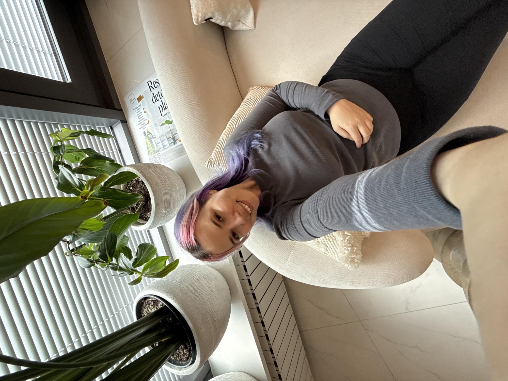 An adult female, with pink and purple hair, is depicted in a selfie within an indoor setting in the City of Zagreb, Croatia. She is positioned on a light-colored, curved sofa, wearing a grey long-sleeved top and black pants, smiling towards the camera. Two decorative pillows, one striped and one textured cream, are present on the sofa. In the background, a window with horizontal blinds is visible, flanked by three potted plants in white planters. A white radiator cover is situated below the window. A crumpled document, featuring a QR code and partially legible Croatian text 'Res detc pla', rests near the window. The floor is tiled with a light, subtly marbled pattern.