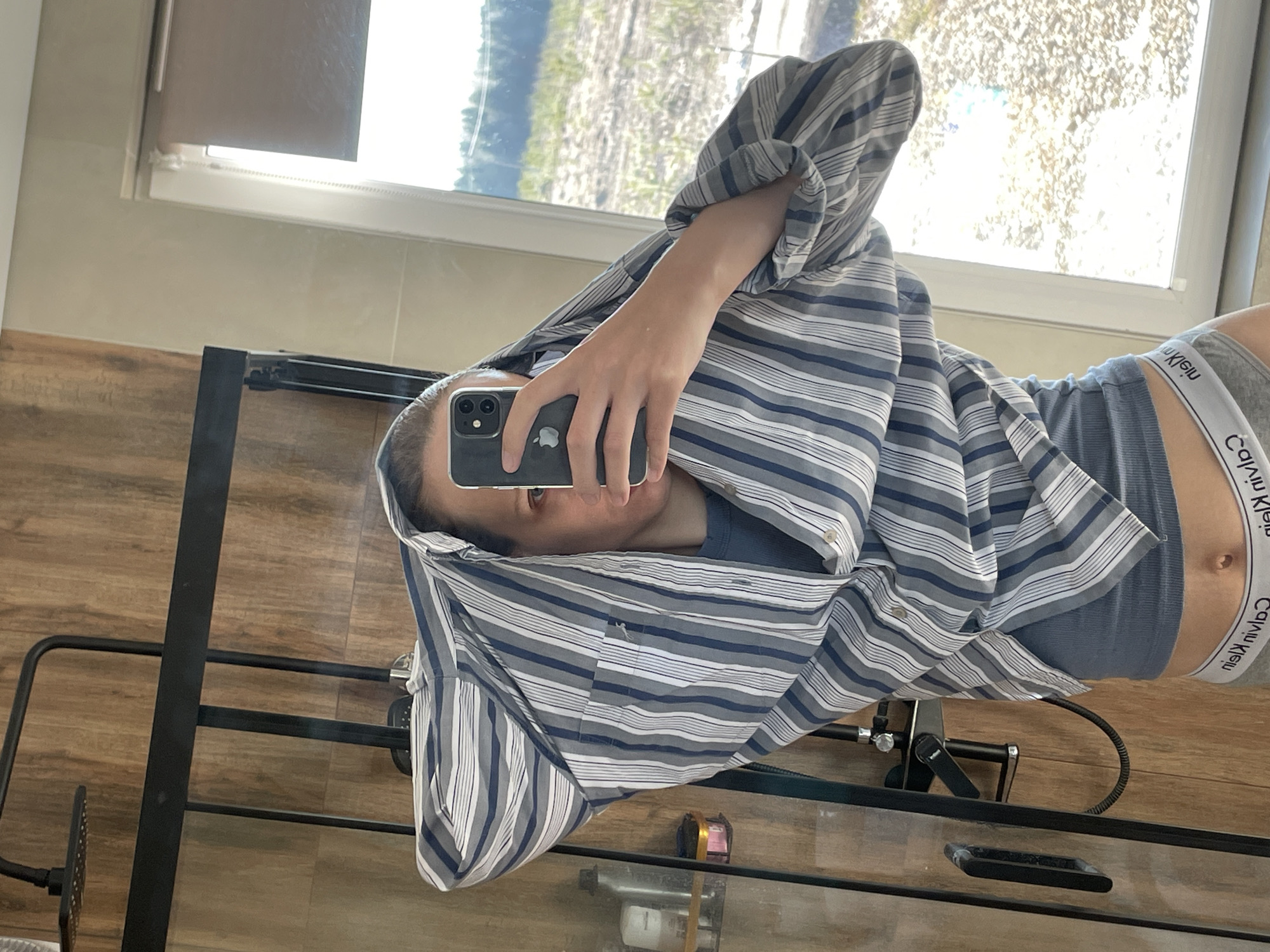 The image shows a person taking a mirror selfie in what appears to be a bathroom. The person is wearing Calvin Klein underwear and a striped shirt, partially covering their face with the shirt. They are holding an iPhone to take the picture. The background shows a window and part of a dresser or table. The image has been rotated. It is difficult to ascertain further details about the location. Therefore, it is not possible to confirm that the picture was taken in Podshivalovo, Russia.