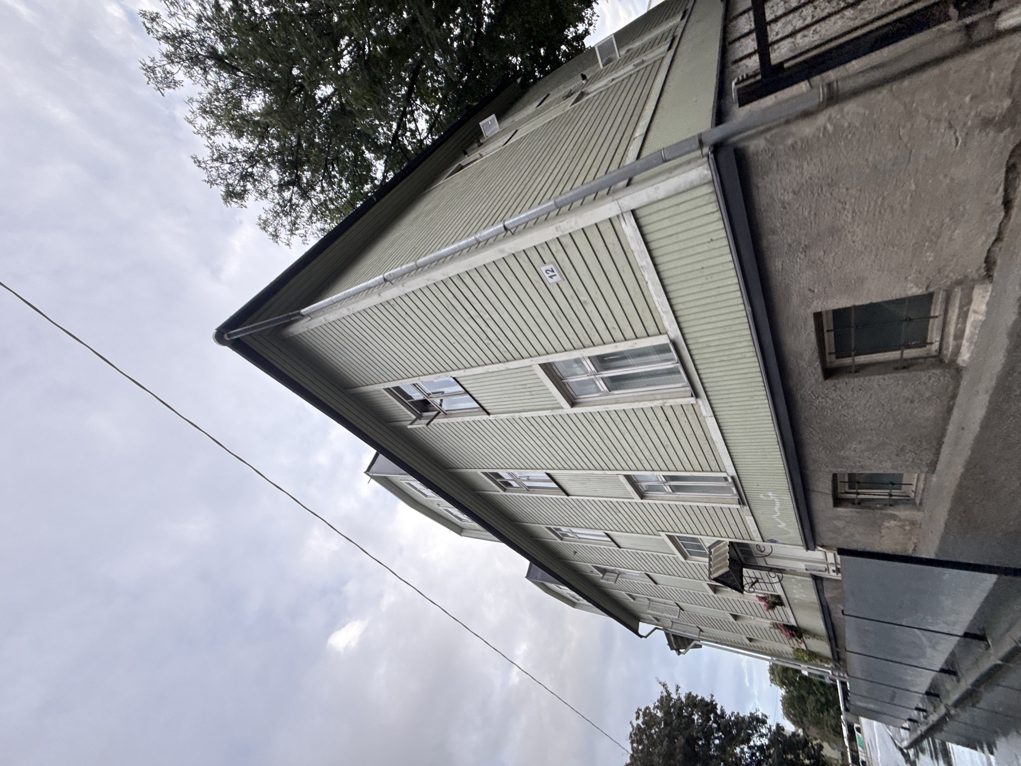 A low-angle, wide shot captures the exterior of a light green, multi-story wooden building in Tallinn, Estonia. The building features multiple windows with white frames, some of which are open. A prominent drainpipe runs down the side of the building, and a small number "12" is affixed to the siding. The building has a stone foundation, and a dark metal fence runs along the front of the property. The sky is overcast and cloudy. To the right of the building, a large tree with dark green foliage is visible. The scene appears to be on a street, with a glimpse of traffic and other buildings in the background.