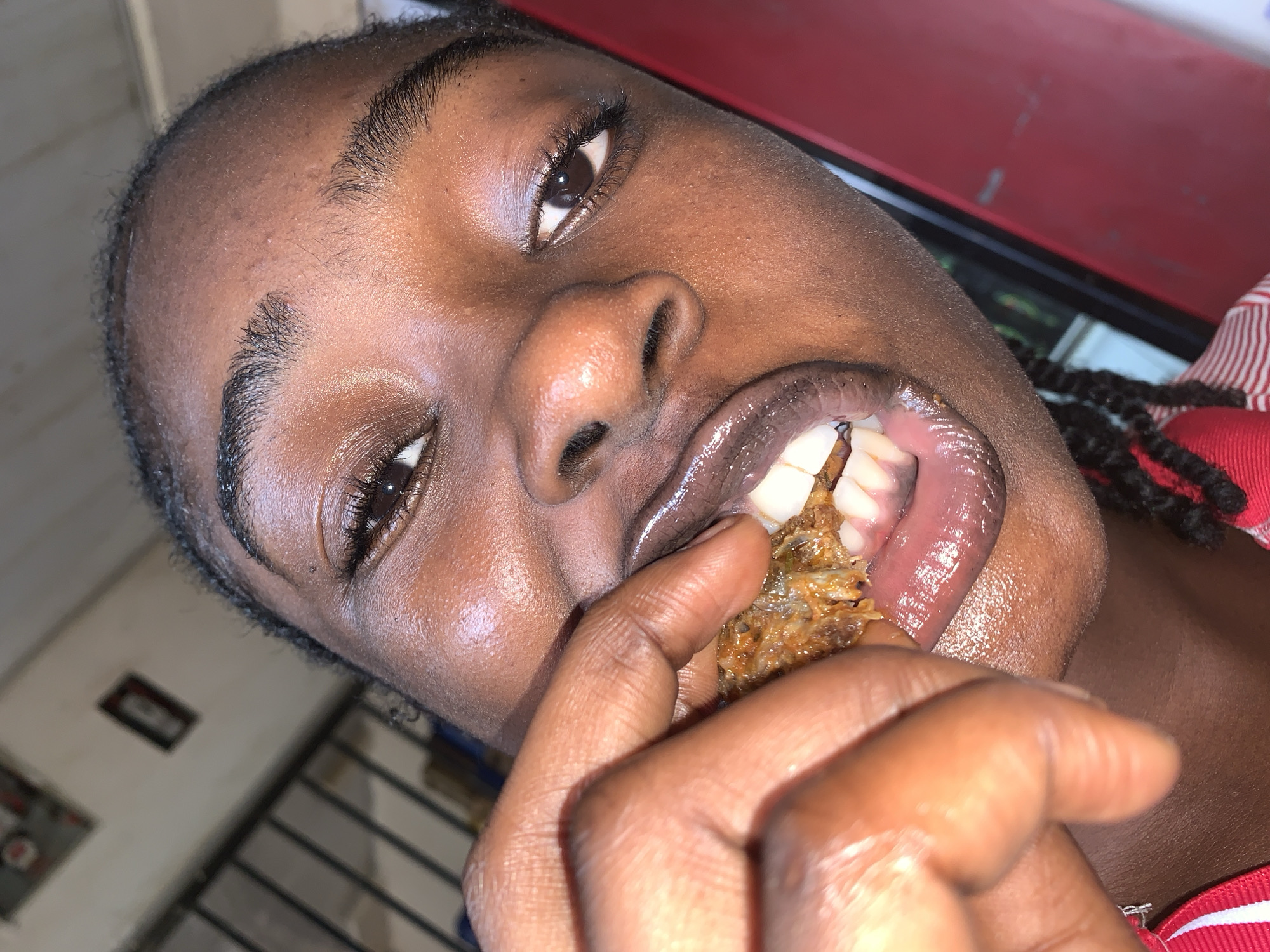 A close-up, angled portrait features a person with dark skin actively eating. The individual's face is prominent, tilted slightly, with their mouth open, revealing visible teeth as they bite into a brown, textured food item held between their fingers. The food appears to be a savory snack, possibly fried fish or a similar item, given its fibrous texture and small pieces.
The person has visible braided hair on the right side of the frame and fine, dark hair along their forehead. Their skin has a natural sheen, particularly on the glossy lips and around the eyes. Their dark eyes look towards the viewer, with distinct eyelashes.
The background is partially visible and blurred. A large, reflective red surface dominates the upper right portion of the frame, possibly a wall or a piece of furniture. To the lower left, a dimly lit interior space can be seen, including what appears to be a framed picture on a wall and horizontal bars or lines, possibly from a window or railing. The person is wearing a red and white striped garment. The lighting suggests an indoor setting, likely in the evening or at night. The activity centers on the immediate act of eating. The scene is situated in New Stadium, Nigeria. No text is visible.