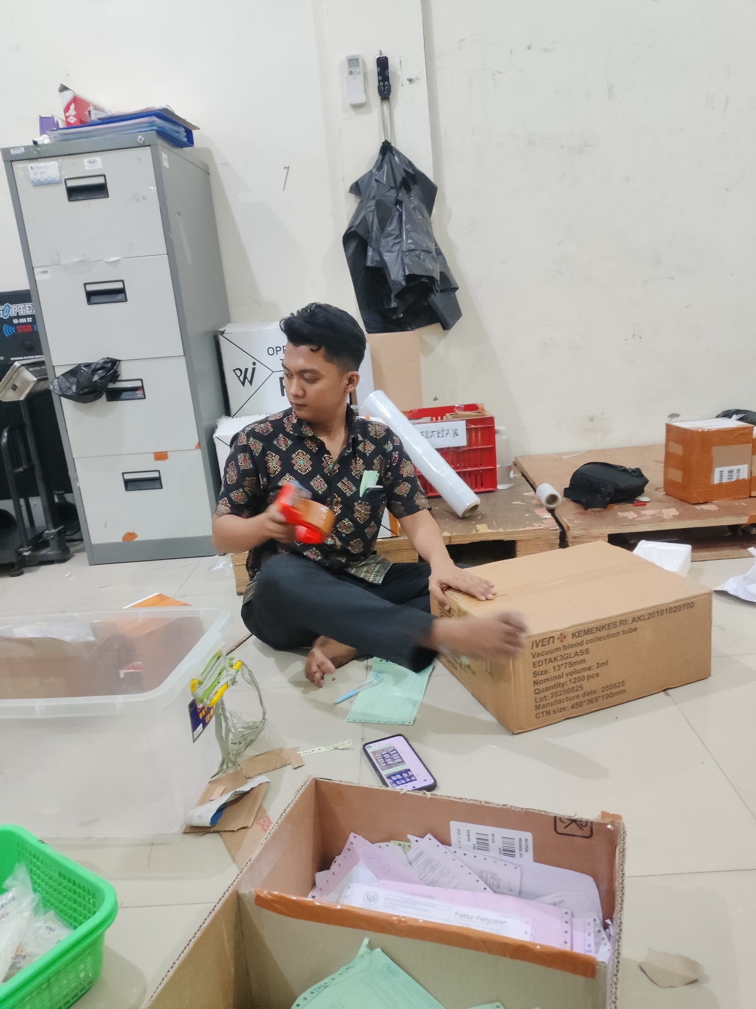 The image depicts an indoor scene, likely a storage or packing area, with a man sitting on the floor surrounded by boxes and other items. The man is wearing a dark patterned shirt and dark pants, and he is holding a small orange container in his right hand, appearing to be examining it. In front of him, a large cardboard box is open, filled with papers, and next to it is a clear plastic container.
To the left, a grey filing cabinet is visible, with some papers and folders on top. There's also a green plastic basket containing white packaged items. In the background, more boxes, rolls of plastic wrap, and red plastic crates are scattered around, suggesting an active work environment. The walls are a light color and appear somewhat worn.
The overall impression is one of a functional workspace where goods are being processed or organized. The presence of medical supply labels on some boxes (e.g., "Vacuum blood collection tube") indicates the nature of the items being handled. The scene suggests a busy, possibly late-day, work session. The specific location of Pepelegi, Indonesia, is noted, indicating a likely distribution or fulfillment center within that area.
