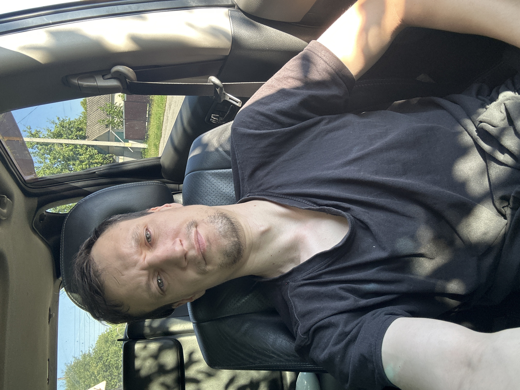 A medium shot captures a man with short, dark hair and light eyes sitting in the driver's seat of a car, facing the camera. He has a short beard and is wearing a dark t-shirt. Sunlight filters through the car window, casting shadows on his face and shirt. The background outside the window shows trees and a building, suggesting a rural or suburban setting. The location is identified as Shatava, Ukraine.