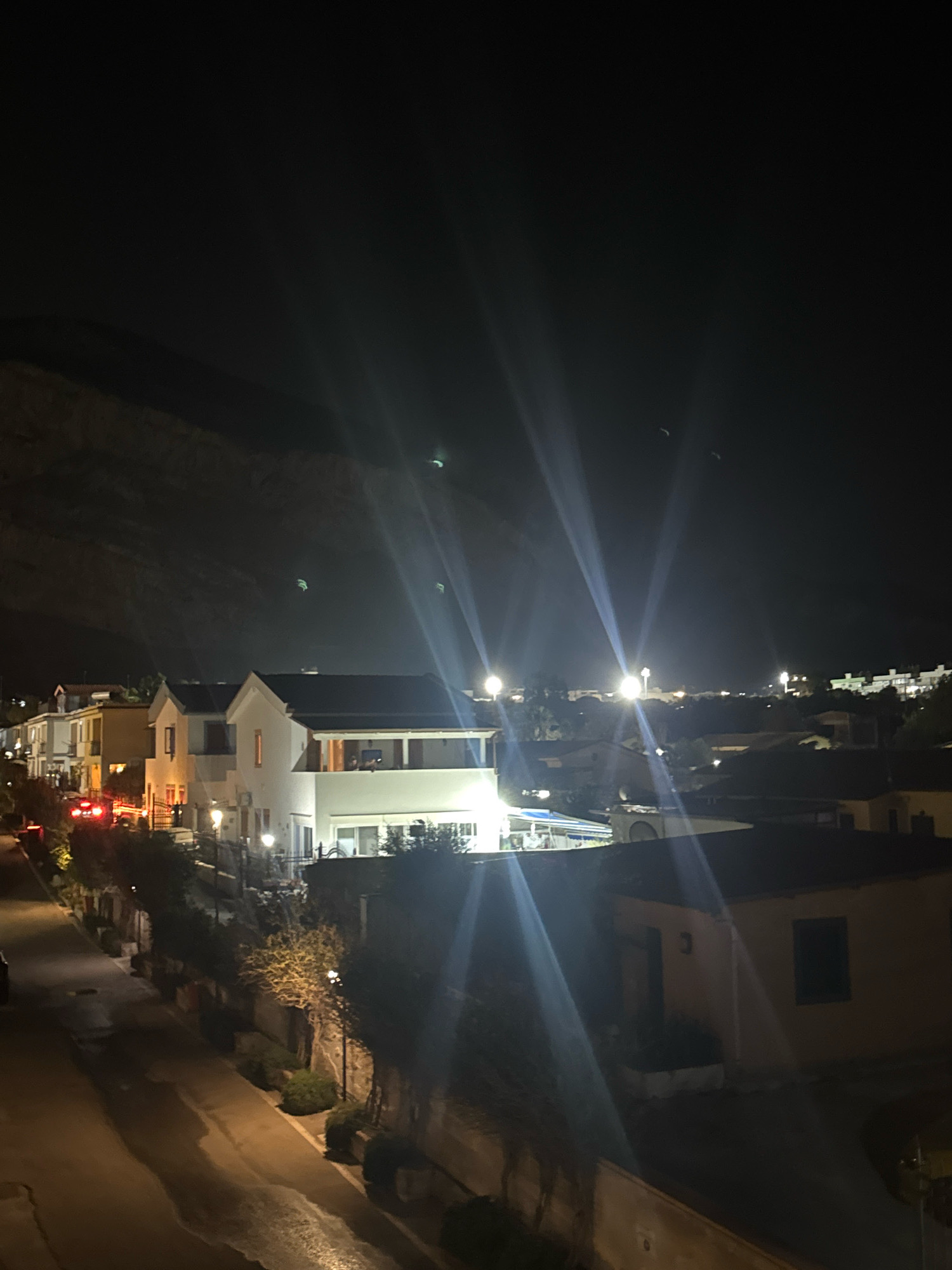 The image is a nighttime photograph taken from an elevated position, overlooking a street with houses and a hillside in the background. The scene is illuminated by artificial lights from streetlamps, house interiors, and vehicle lights. Several houses are visible, appearing to be residential buildings with varying architectural styles, some with balconies and illuminated windows. A street runs through the foreground, with a wet surface indicating recent rain. Red taillights of vehicles are visible on the street, suggesting traffic. The sky is dark, with some light streaks caused by lens flare from bright light sources. In the upper part of the image, several faint green lights are visible, possibly drones or lanterns. The location is identified as Capaci, Italy.