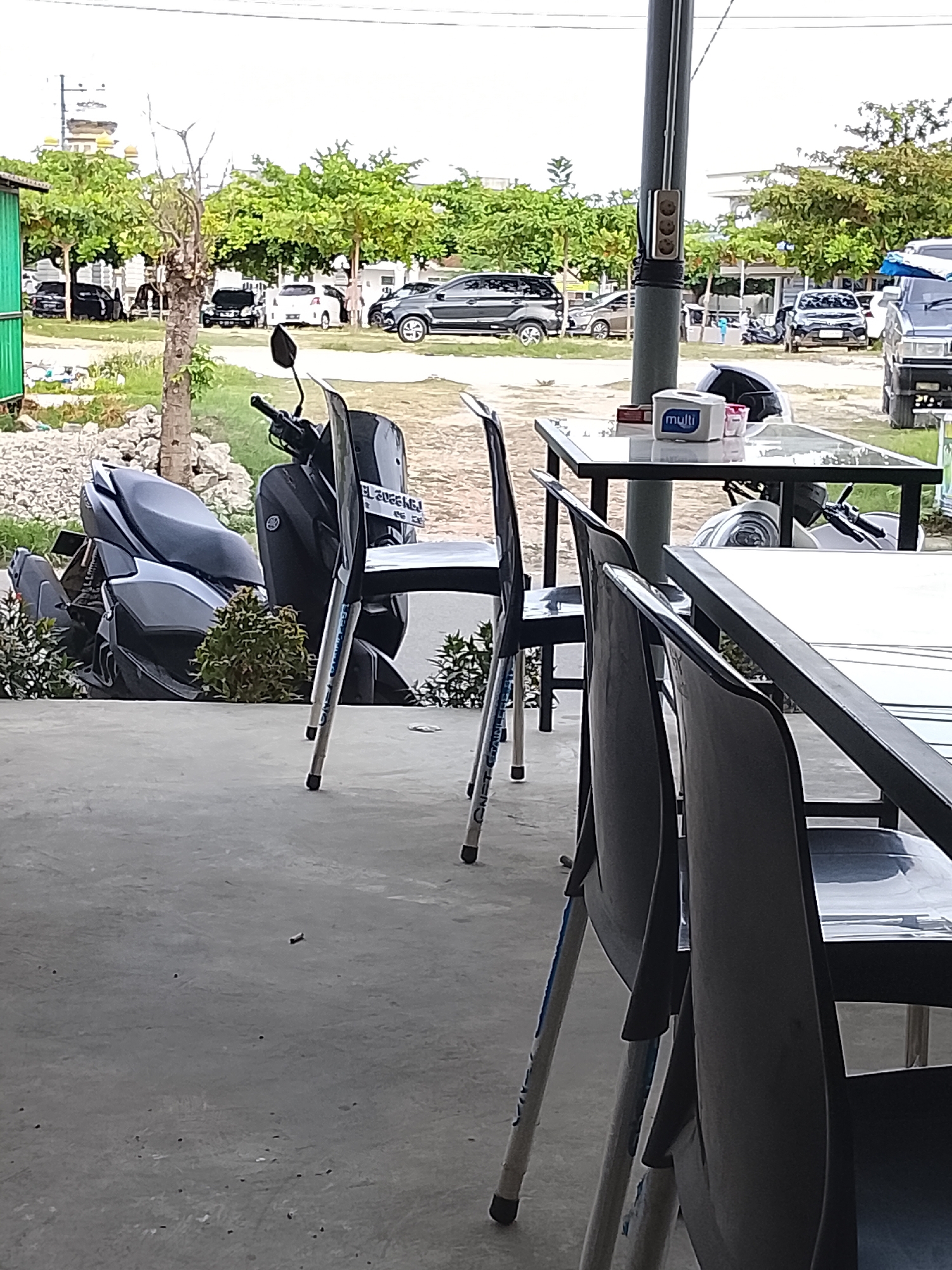 The image shows a cafe setting in Lhokseumawe, Indonesia. The foreground features tables and chairs. A dark-colored motorcycle is visible behind a chair, and a parking area with several cars is in the background. The sky is overcast.