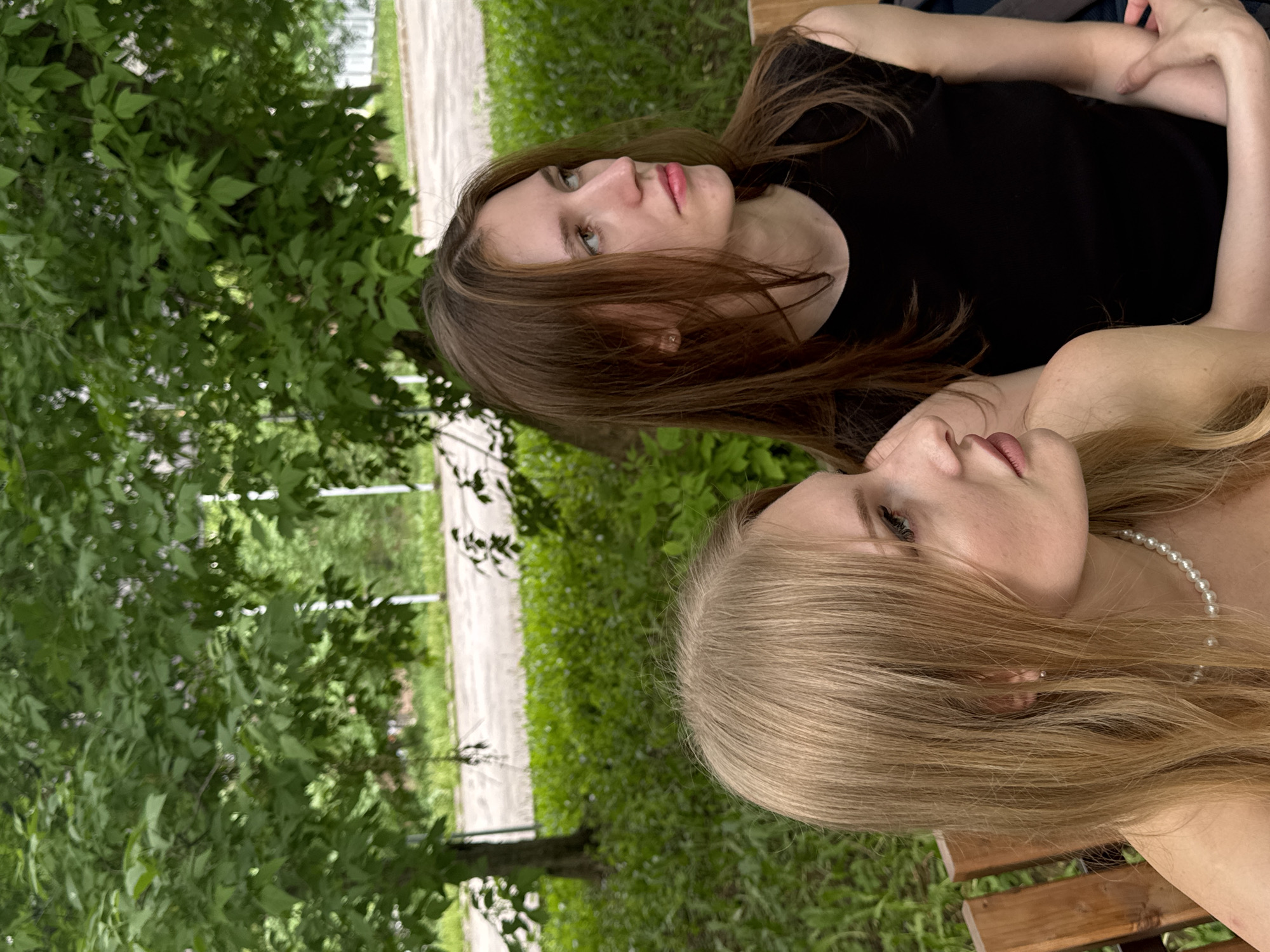 Two young women are lying on a wooden bench outdoors. The woman closer to the viewer has long blonde hair and is wearing a pearl necklace. The other woman has long brown hair and is wearing a black top. They are lying next to each other, looking in different directions. The background includes green foliage, grass, and a paved path. The location appears to be a park or garden. This is Perm, Russia.