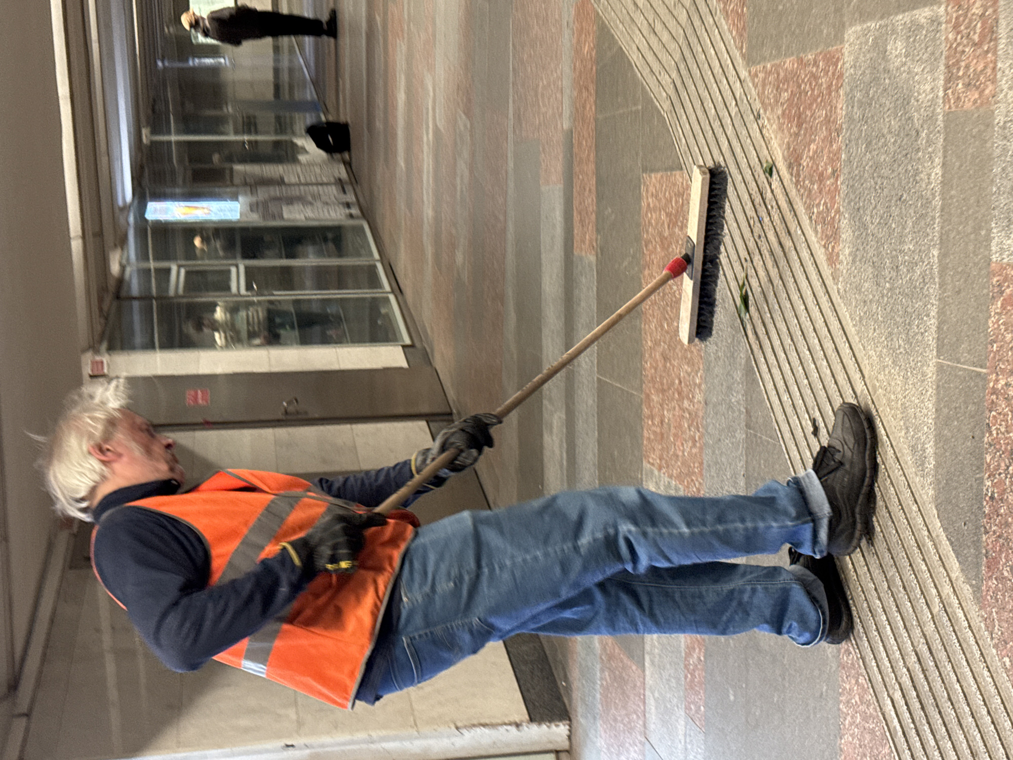 A man with messy blonde hair is sweeping the floor in a covered walkway, possibly a metro station. He is wearing a dark blue long-sleeved shirt, a bright orange safety vest with reflective stripes, blue jeans, and black gloves. He is holding a wooden broom with a red ferrule and a black brush head. The floor is made of patterned tiles with pink and grey sections. There are raised tactile paving strips running diagonally across the floor. In the background, glass storefronts line the walkway, and other people are visible, including a person wearing a light-colored cap in the distance. The environment appears to be indoors, with artificial lighting. The image is taken from a low angle, focusing on the man and his sweeping action.