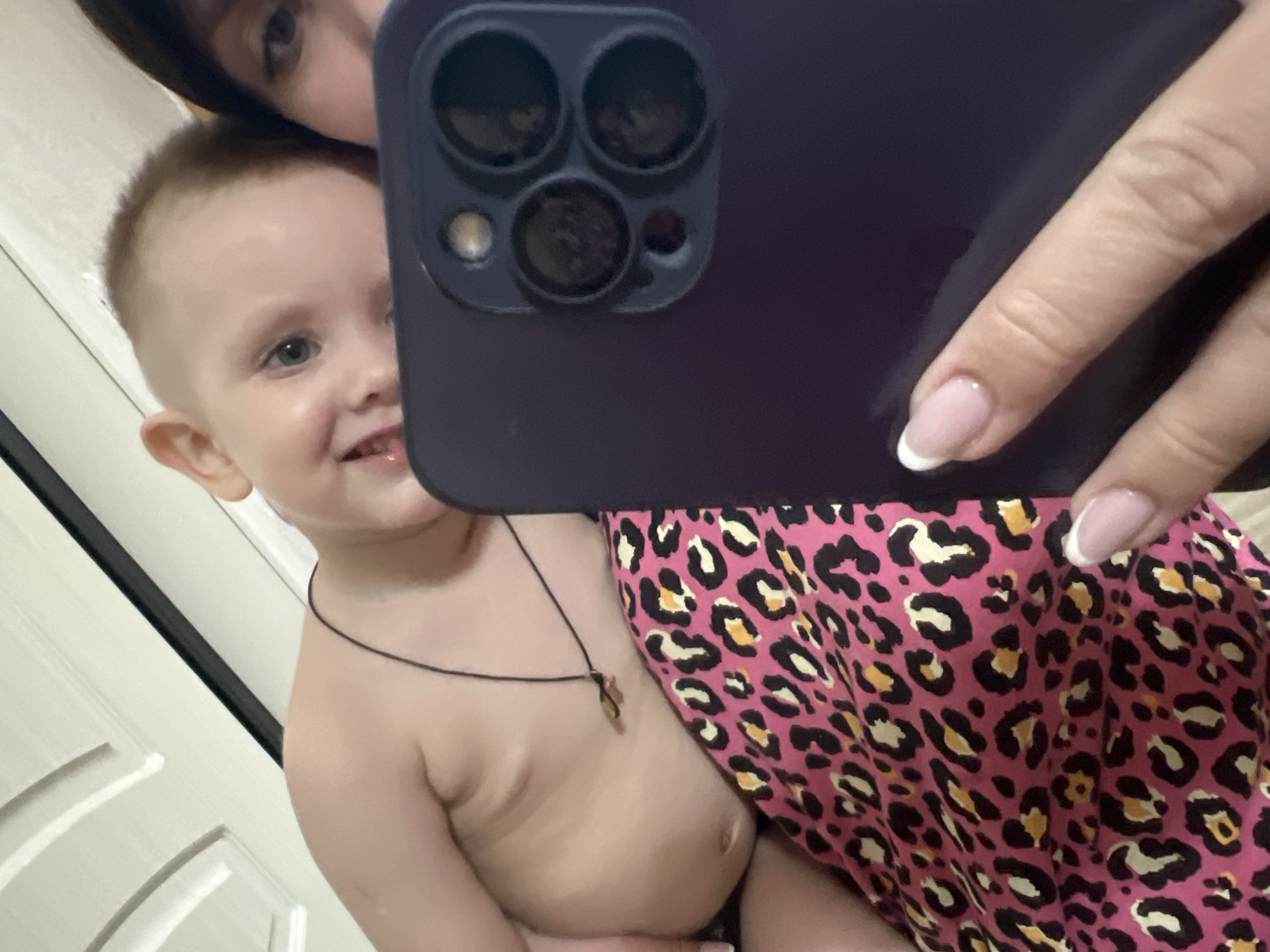 A woman is holding a smartphone to take a selfie with a young child. The phone, dark purple, is an iPhone with three lenses. The woman's right hand is holding the phone, and her fingers have light pink nails with white tips. The child, likely a toddler, has light skin, a short haircut, and is wearing a thin necklace with a cross. The child is partially undressed. The woman is partially visible above the phone. A pink and black leopard-print garment covers the lower part of the frame. A white door with molded panels is partially visible in the background. It is not possible to verify the precise location of Марковский, Russia based solely on the visual data.
