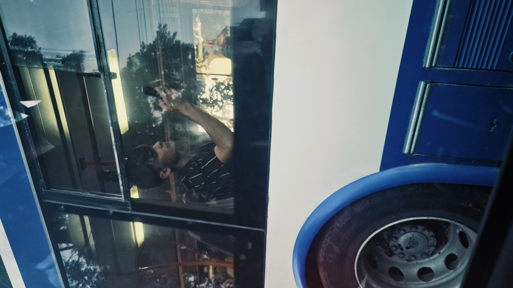 A young man with dark hair, wearing a black and white striped t-shirt, is visible through the window of a bus. He is holding a smartphone in his right hand, seemingly taking a picture or recording a video, with his arm extended towards the window. The bus appears to be in motion, as evidenced by the blurred reflection of trees and the sky in the window. The exterior of the bus is white with a blue stripe along the bottom, and a portion of a large tire is visible in the foreground on the left. The scene suggests an everyday moment captured during a bus journey in Chișinău, Moldova.