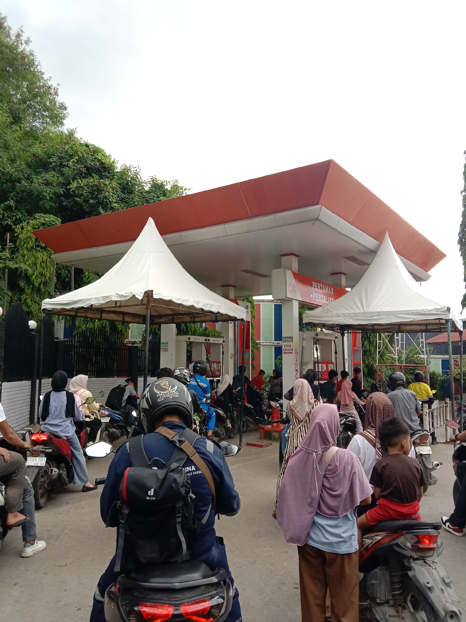 The image depicts a busy scene at a gas station in Lhokseumawe, Indonesia. A large group of people on motorcycles are lined up, presumably waiting to purchase fuel. The gas station has a red and white awning and is also outfitted with two white tents on each side of the entrance, presumably for shade. The sky overhead is overcast and the surrounding area features green trees and glimpses of other buildings. Several brand names for gasoline (PERTAMAX, PERTALITE) are visible. In the foreground, people are mostly on motorbikes, with a few standing on the ground.