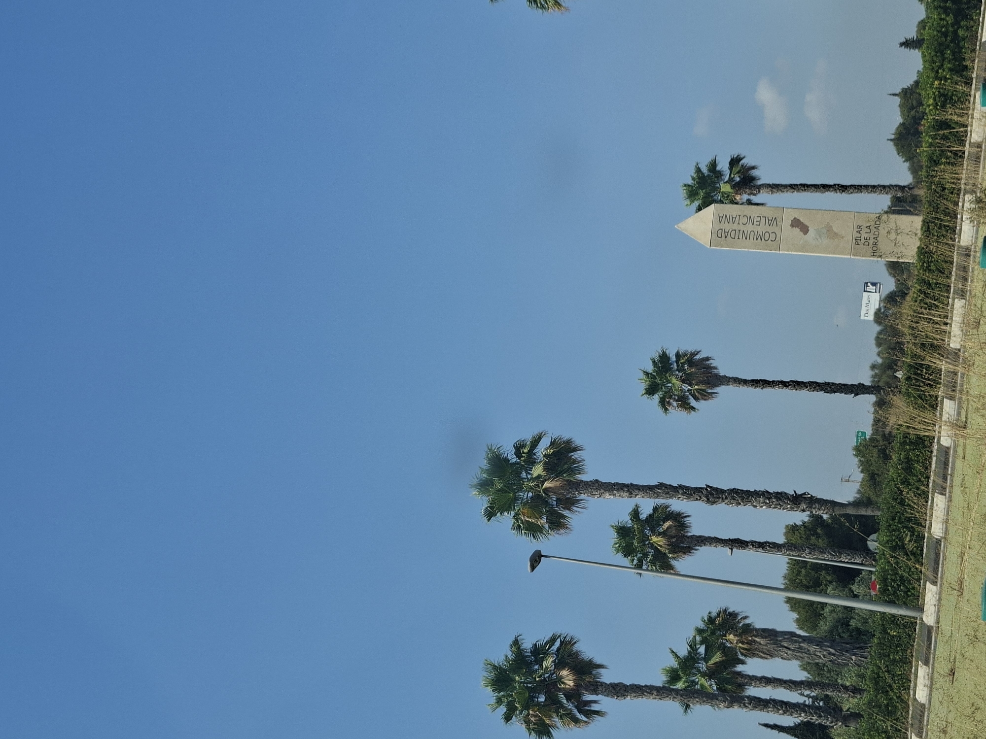 The image shows a bright, sunny day with a clear blue sky above. A row of tall palm trees stands against the sky on the right side of the frame. In the foreground, a beige, obelisk-shaped monument is visible, inscribed with text that reads "COMUNIDAD VALENCIANA" and "PILAR DE LA HORADADA". The monument appears to be situated in an outdoor area with dense green bushes and vegetation. To the left of the monument, a street lamp is positioned between two palm trees. A sign for "Dos Mares" is also faintly visible behind the monument. The overall scene suggests an exterior location in the town of Pilar de la Horadada, Spain, likely a roadside or public area.