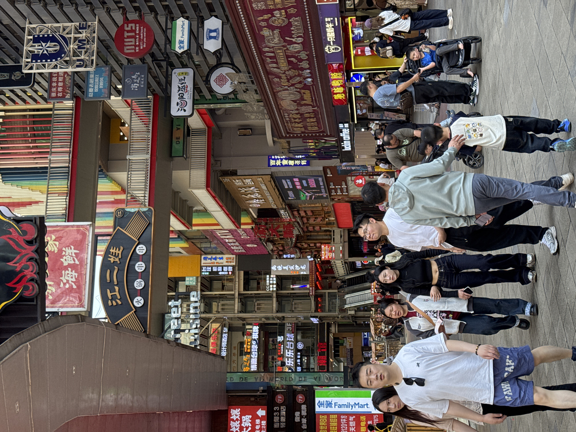 The image captures a street scene in Yuzhong District, China, with numerous shops and restaurants indicated by various signs in Chinese and English. A mix of people, including groups of friends, are visible walking along the street. The signs overhead and on buildings display a diverse range of businesses, including food establishments, entertainment venues, and hotels. The street appears bustling with activity, as evidenced by the number of pedestrians. The image's composition gives a sense of depth with the buildings and signs receding into the distance. Notable brands such as FamilyMart and Luckin Coffee are also present.
