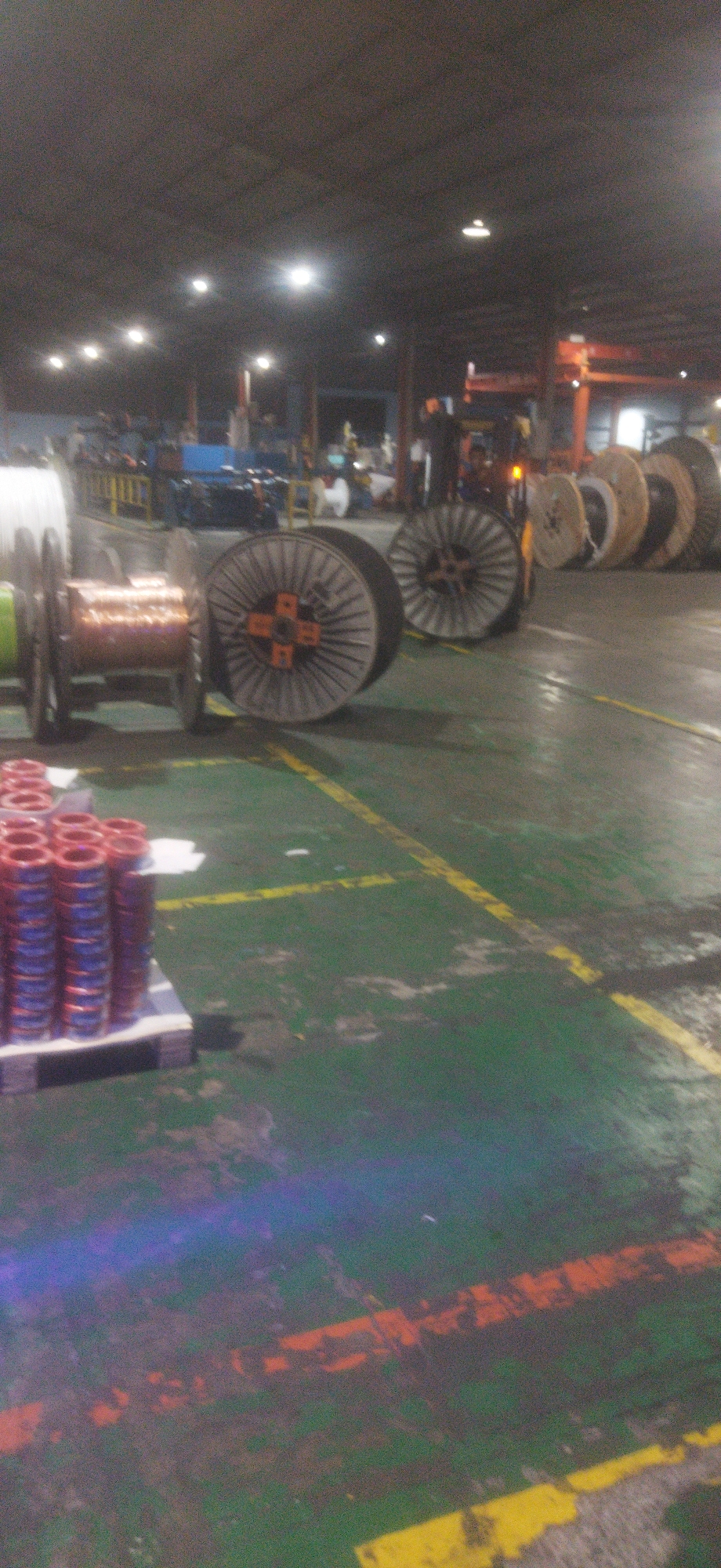 The image depicts the interior of a manufacturing facility in Cileungsi, Indonesia. The environment is a large, open industrial space with a high ceiling and visible overhead lighting. The floor is painted green with yellow and orange safety markings. In the foreground, a pallet loaded with spools of red and blue wire is visible on the left. To the right of the pallet are several large industrial spools, some appearing to be made of wood and metal. One prominent spool in the mid-ground has a copper-colored winding. In the background, more industrial spools are lined up, and various machinery and equipment associated with manufacturing are present. The overall impression is a busy production or storage area.