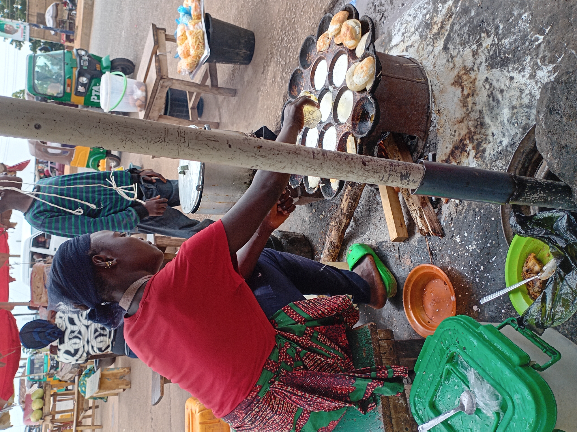 A street scene in Bukuru, Nigeria, shows a woman in a red shirt and patterned skirt sitting on a low stool, cooking small, round cakes in a multi-compartment iron pan over a wood fire. She pours oil from a clear bottle into one of the cooking compartments. A man in a plaid shirt sits in the background. Other street vendors sell fried snacks from wooden stands. A green auto rickshaw is parked nearby. A green lunchbox, an orange bowl, and a green plate with food are visible on the ground near the woman. A metal bar is visible in the foreground.