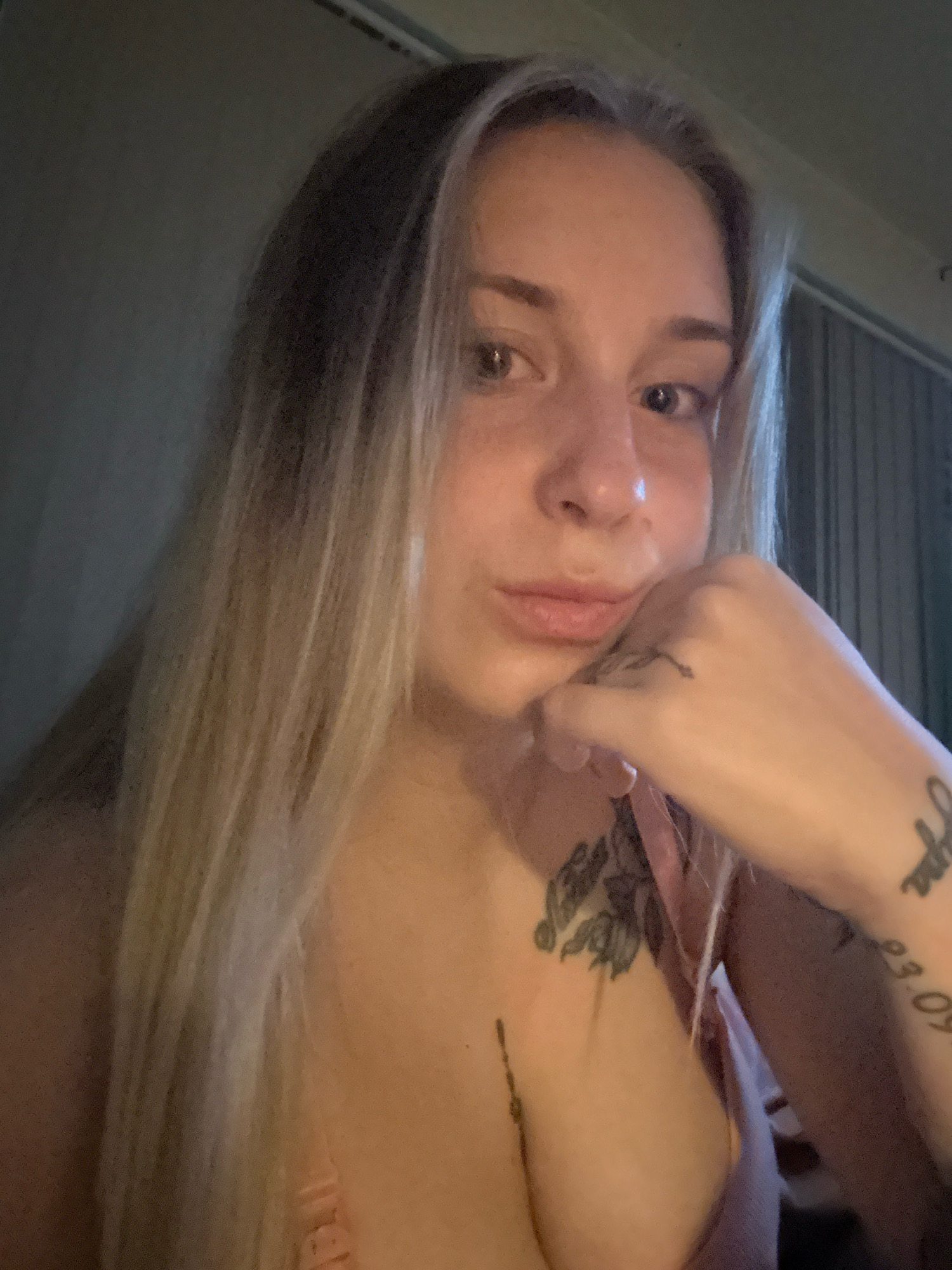 A close-up, chest-up, indoor selfie shows a woman with long, blonde hair and light skin. She is resting her chin on her hand, which has a tattoo on the back of it. She also has a tattoo on her chest. The background appears to be a room with vertical blinds. The location of this image is not Plovdiv, Bulgaria; it is an unknown indoor location.