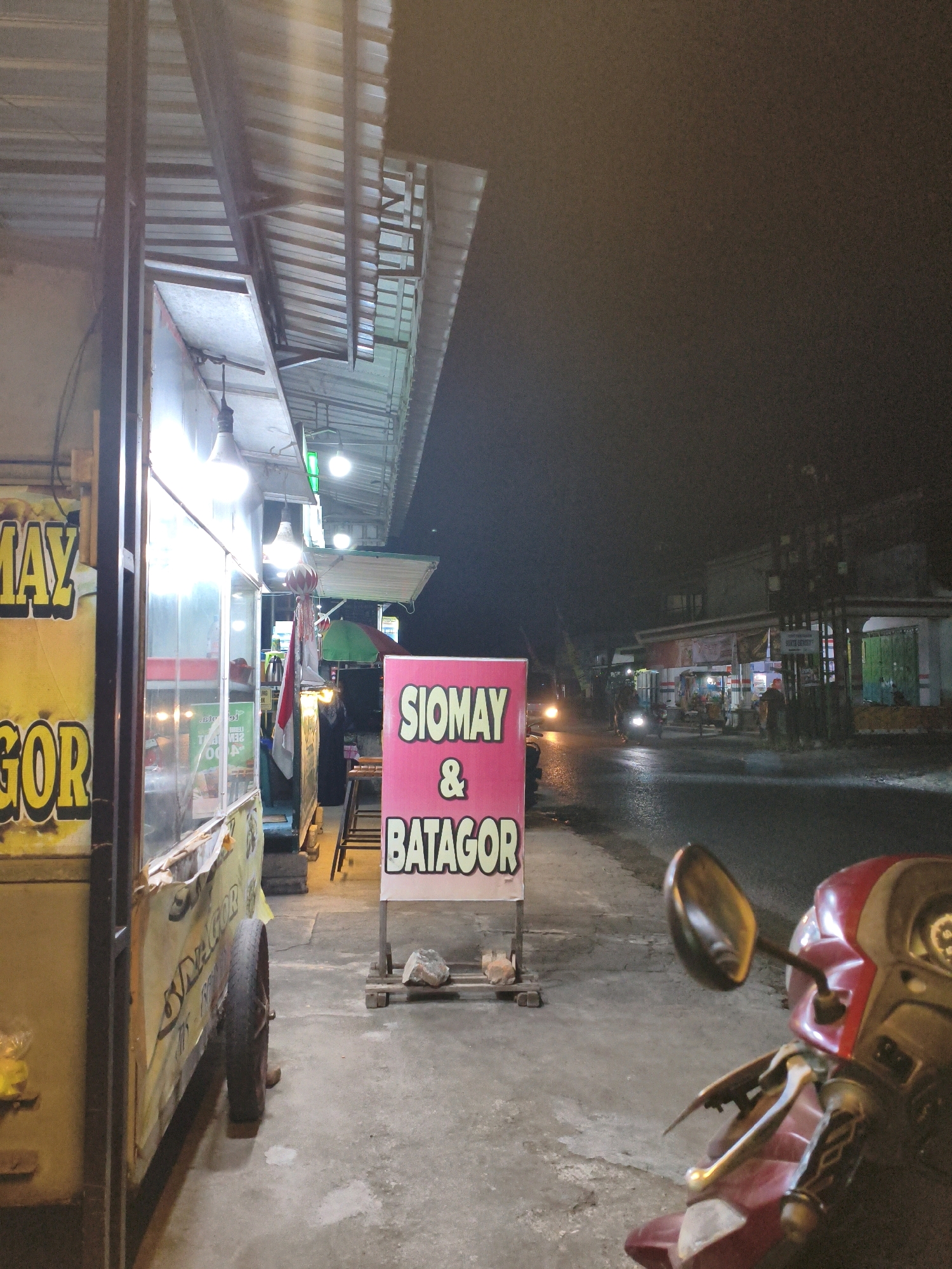 A night scene depicts a street-side food stall in Tunggangri, Indonesia. A food cart, predominantly yellow with large lettering partially visible, is on the left. To its right, a pink sign displays "SIOMAY & BATAGOR" in white text. The street is wet and reflects the lights of passing vehicles and street lamps. A red scooter is parked in the foreground on the right. Buildings line the street in the background, and several people are visible near other stalls or walking. The sky is dark and overcast.