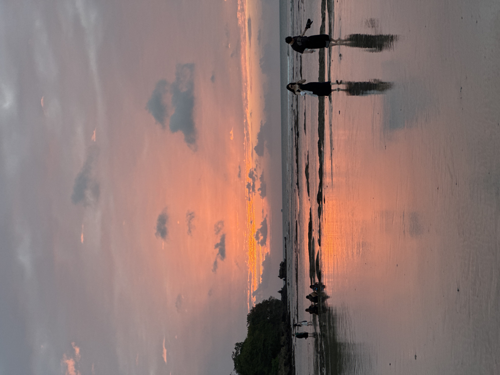 The image depicts a serene beach scene during what appears to be sunset or sunrise at the coast of Kuta Selatan, Indonesia. The sky is awash in soft hues of pink and orange, with scattered clouds reflecting the warm light. The ocean is calm, with gentle waves lapping at the shore. In the foreground, a group of people are visible, some standing and others sitting on the wet sand. Their reflections are mirrored in the shallow water covering the beach. In the mid-ground, two individuals, a woman in a black dress and a man in dark clothing, are walking along the water's edge. Further in the background, a lush green tree-covered cliff juts out into the sea, adding a touch of natural beauty to the landscape. The overall atmosphere is tranquil and picturesque, capturing the peaceful ambiance of a tropical beach at twilight.