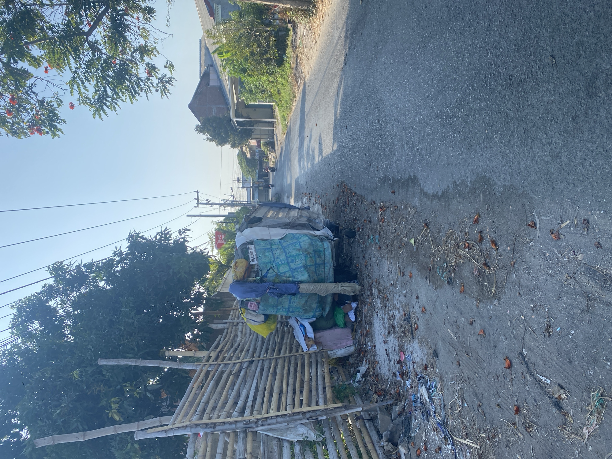 The image depicts a street scene in Kediri City, Indonesia. On the left side of the paved road, there is a bamboo fence and a cart loaded with various items, covered with a blue tarp. A person, partially visible, is interacting with the cart. The ground is littered with debris, including dry leaves and trash. Beyond the cart, there are trees and foliage. The road continues into the distance, with houses and more vegetation visible on the right side. Power lines run overhead. The sky is clear and blue, indicating daylight.