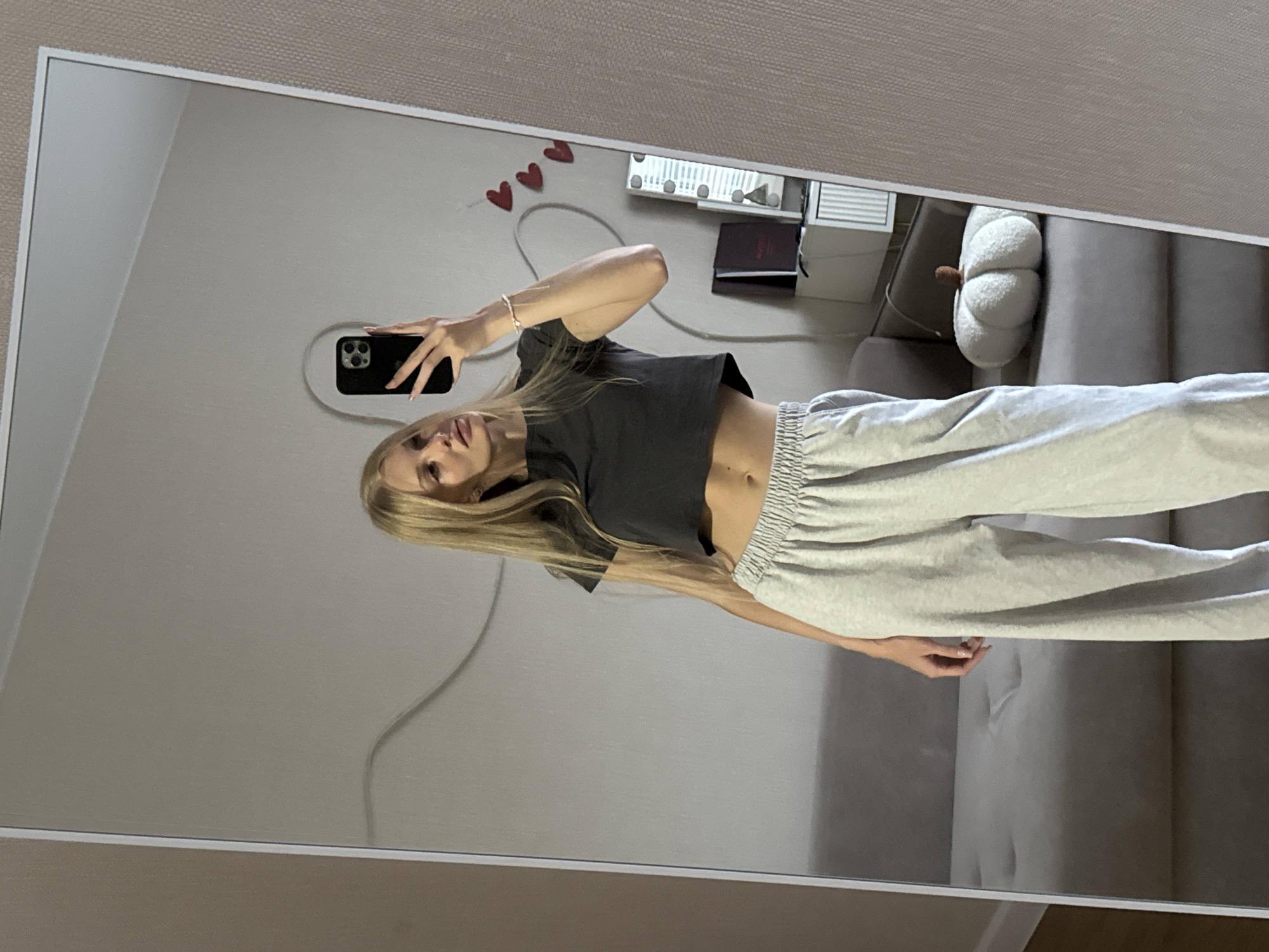 A young woman with blonde hair is taking a mirror selfie in an indoor setting. She is wearing a dark grey crop top and light grey sweatpants. Her left hand is holding a black smartphone, and her right hand is resting on her hip. In the reflection of the mirror, a couch, a white pumpkin-shaped pillow, and some shelves with decorative items are visible. The overall scene appears to be a bedroom or living area. There is no indication of the location being in the city of Perm, Russia.