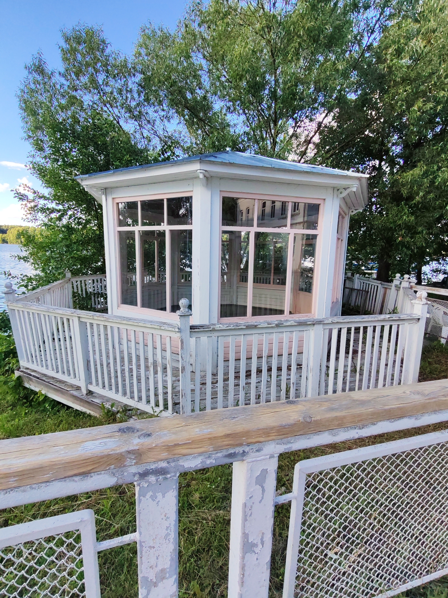 The image depicts a quaint, octagonal gazebo situated on a grassy knoll overlooking a body of water, likely a river or lake, in Plyos, Russia. The gazebo is painted white with pale pink trim around its large, multi-paned windows. A white wooden railing encircles the gazebo, with decorative balusters and finials. The wood of the railing shows signs of age and wear, with peeling paint. The gazebo is surrounded by lush green trees, some of which are quite large and cast dappled shadows. The sky is a clear, bright blue with a few wispy clouds, suggesting a pleasant day, likely late morning or afternoon given the lighting. In the foreground, a more modern, lower fence with a weathered wooden top rail and wire mesh inserts partially obscures the view of the gazebo and its surrounding railing. The overall impression is one of a serene, somewhat rustic, waterfront setting, possibly a park or a historical estate. There is no text visible in the image.