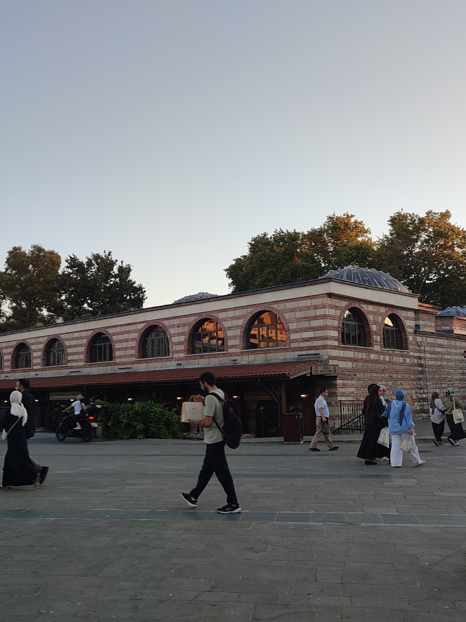 In Üsküdar, Turkey, a group of people are walking on a paved plaza in front of a historic building. The building has a red and white striped facade with arched windows and a dome. Some individuals are dressed in traditional Islamic clothing, including hijabs and long robes. Others are wearing Western-style clothing. Several people are carrying bags. A motorcycle with a passenger is visible in the background. The sky is clear and the lighting suggests it is late afternoon or early evening. Trees with lush green foliage are behind the building.