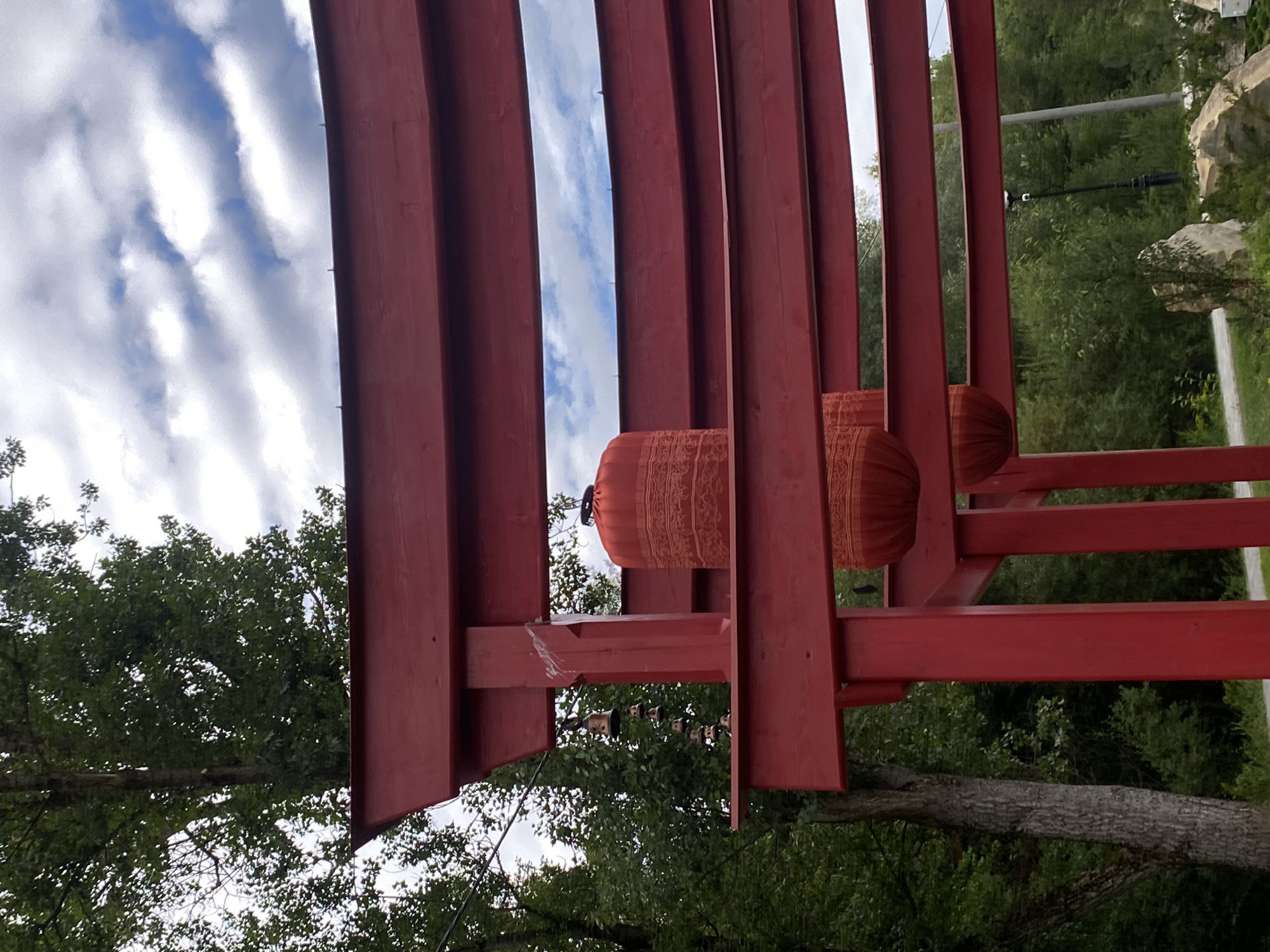 The image shows a red wooden torii gate structure with horizontal beams. Two red lanterns are hanging from the beams, one above the other. The top lantern is cylindrical with intricate patterns, and the bottom lantern appears to be a similar shape. To the left of the torii gate, a string of small, dark bells hangs from a wire. The background consists of lush green trees, a cloudy sky, and some rocks and a lamppost in the distance, suggesting an outdoor, possibly park-like setting. The scene is located in Muszyna, Poland, specifically within the Muszyna Japanese Garden.