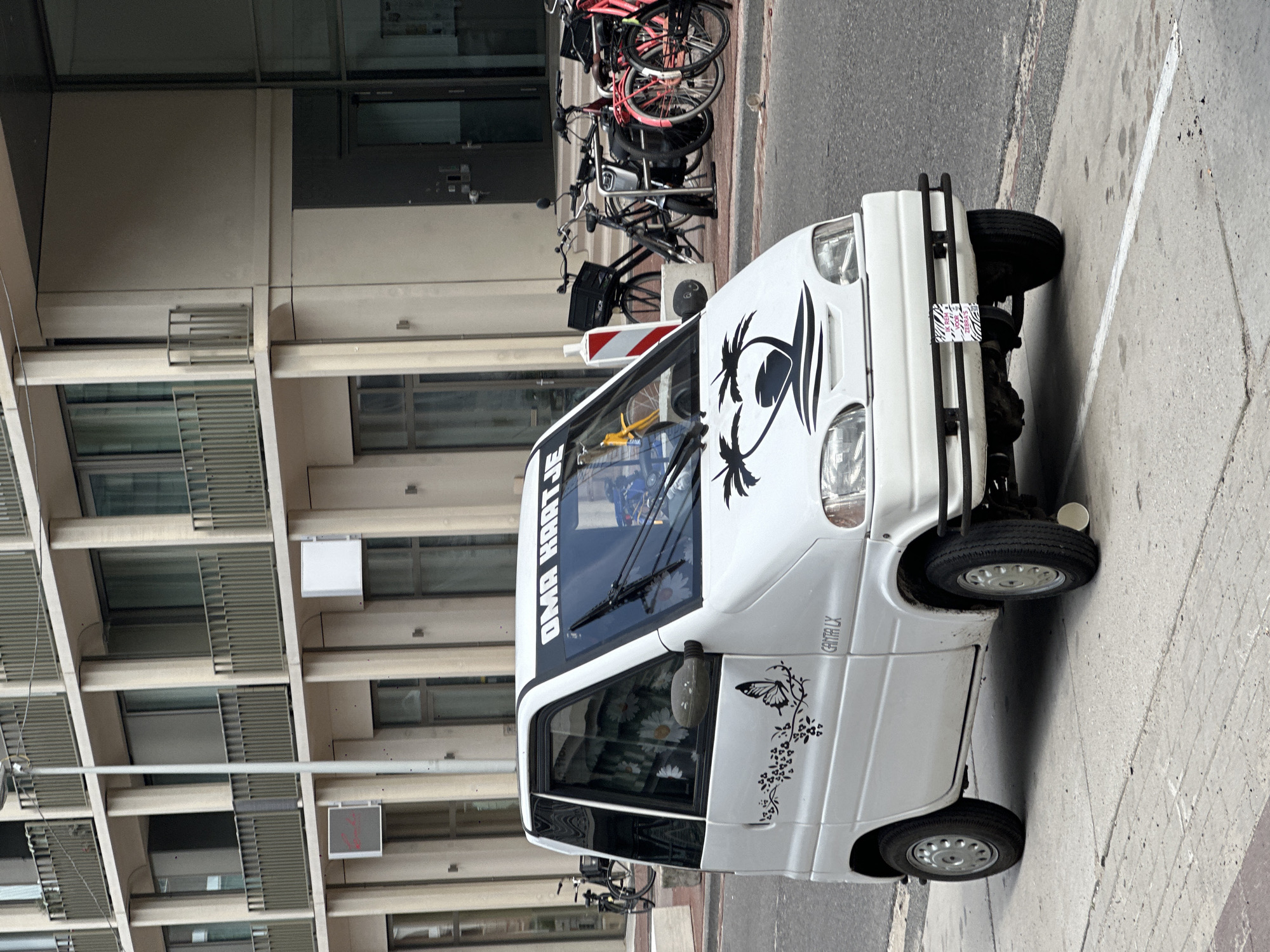 A white, three-wheeled microcar, branded "Canta LX", is parked on a sidewalk next to a street in Amsterdam, Netherlands. The car is decorated with black decals, including a butterfly on the driver's side door and a palm tree silhouette on the hood. A sticker with a zebra pattern and text reading "Ik Ben TESS" is attached to the front bumper. Behind the car, several bicycles are parked along a building. The building itself is modern, with multiple floors featuring balconies and large windows. The street is paved, and the sidewalk is made of concrete slabs with painted white lines. The sky is overcast.