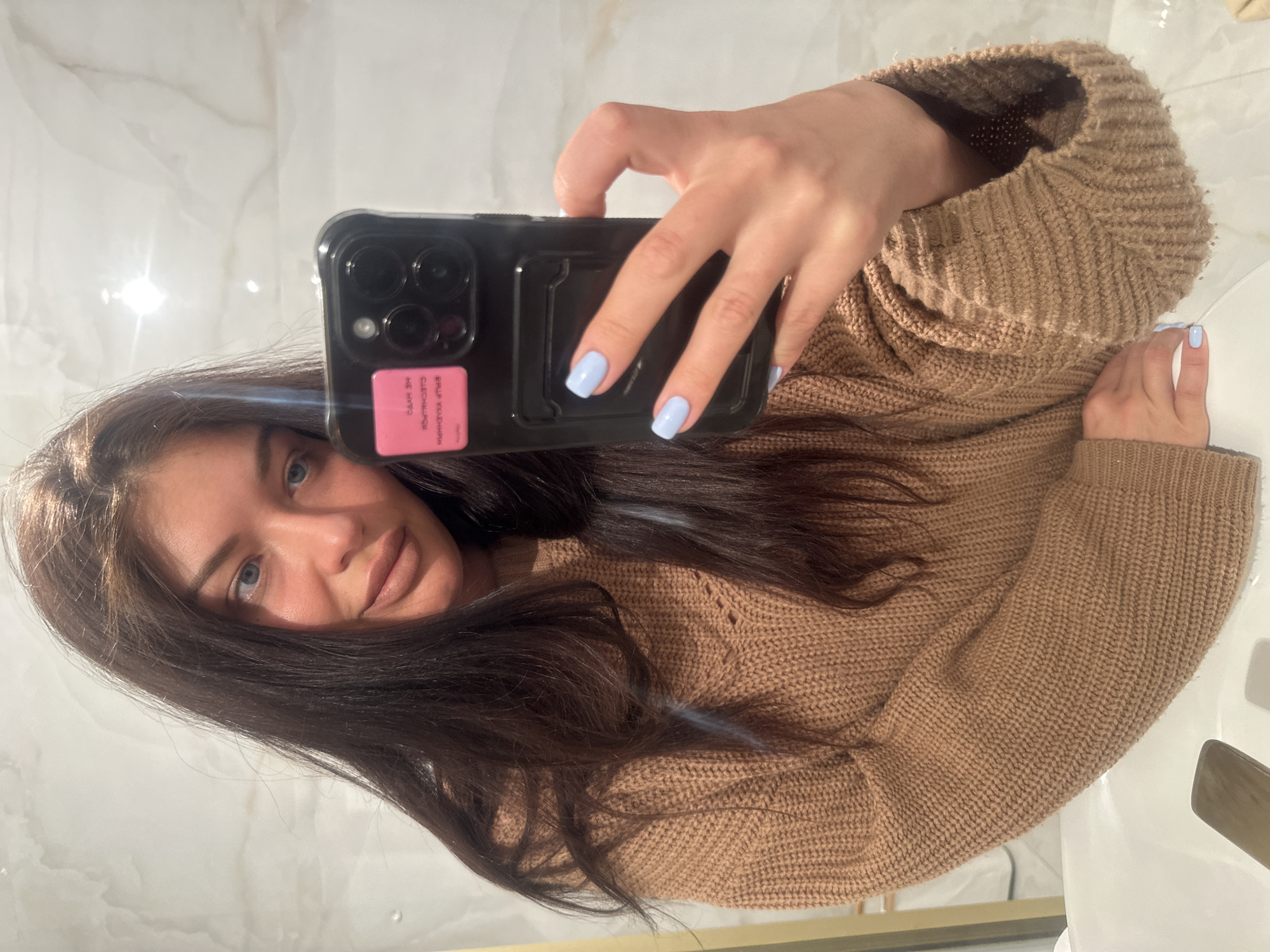 A young woman is taking a selfie with a black iPhone in front of a mirror. She is wearing a brown knitted sweater and has light blue nail polish. The background appears to be a marble tiled wall, possibly in a bathroom. The image was taken in Khimki, Russia.