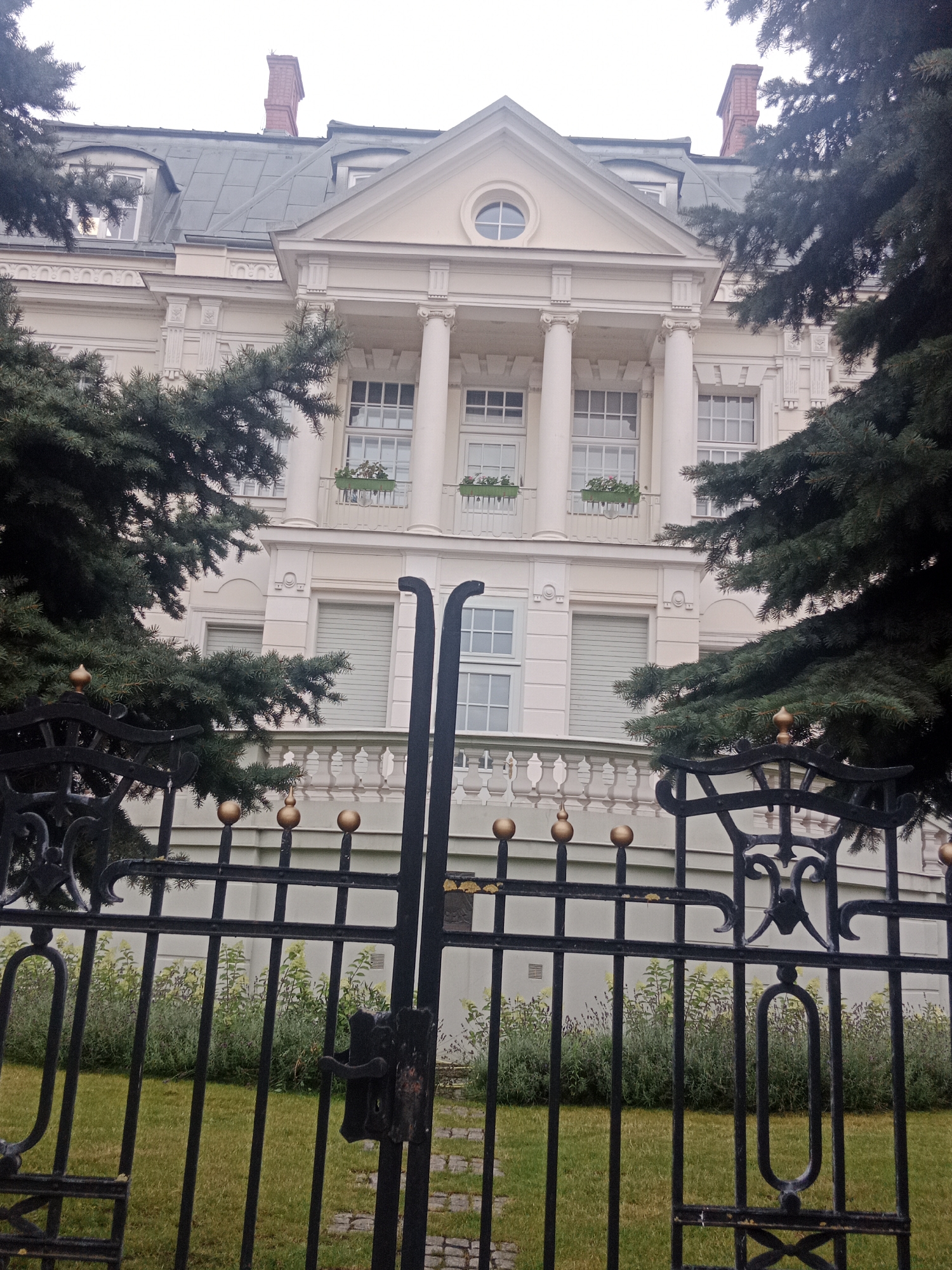 This is a medium shot, vertical, outdoor photograph of a grand, cream-colored building with a grey roof, seen through a black wrought-iron gate and pine trees. The building features classical architectural elements, including a pediment supported by large white columns, multiple windows with white frames, and decorative moldings. A red brick chimney is visible on the roof. Flower boxes with pink flowers adorn the windows on the first level. The gate in the foreground has ornate scrollwork and gold-colored spherical finials. Behind the gate, a well-maintained lawn and a lush garden with green and light yellow foliage are visible. The overall impression is of a stately home or institution, possibly in a park-like setting. The lighting suggests it is daytime, and the overcast sky indicates a cloudy or possibly rainy day. There is no visible text in the image.