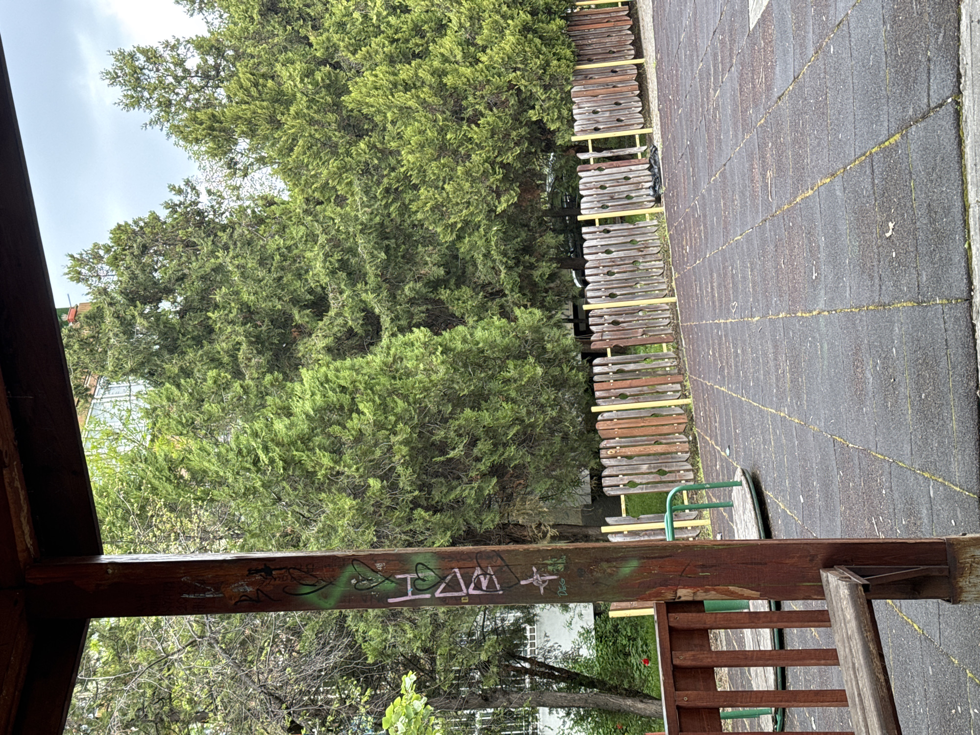 This media file captures an outdoor scene, likely a park or playground in Sofia, Bulgaria, under an overcast sky during daytime. The image, when correctly oriented, presents a vibrant green evergreen tree, possibly a conifer, on the left side, its dense foliage reaching towards the soft light of the sky.
A long wooden structure, resembling a fence or a series of benches with dark wooden slats and bright yellow supports, runs horizontally across the middle ground. To its right, a dark gray paved area, characteristic of a playground surface, extends across the frame. This surface is divided into rectangular sections by yellow lines or strips of moss.
In the foreground, a prominent dark wooden beam stands vertically, showing age and wear. It is adorned with graffiti, including the phrase "I AM" (or a similar stylized word) in pinkish-purple spray paint, along with green spray paint marks and other smaller tags like "DEG" or "DEC." This beam is part of a larger wooden structure, potentially a pergola or part of a playground set, with additional wooden railings and a green metal bar visible at its base and further into the scene. A partial view of a round, green metal structure, possibly a merry-go-round base, is also visible near the paved area.
The scene is devoid of people, conveying a quiet and still atmosphere, highlighting the blend of natural growth and man-made recreational structures.