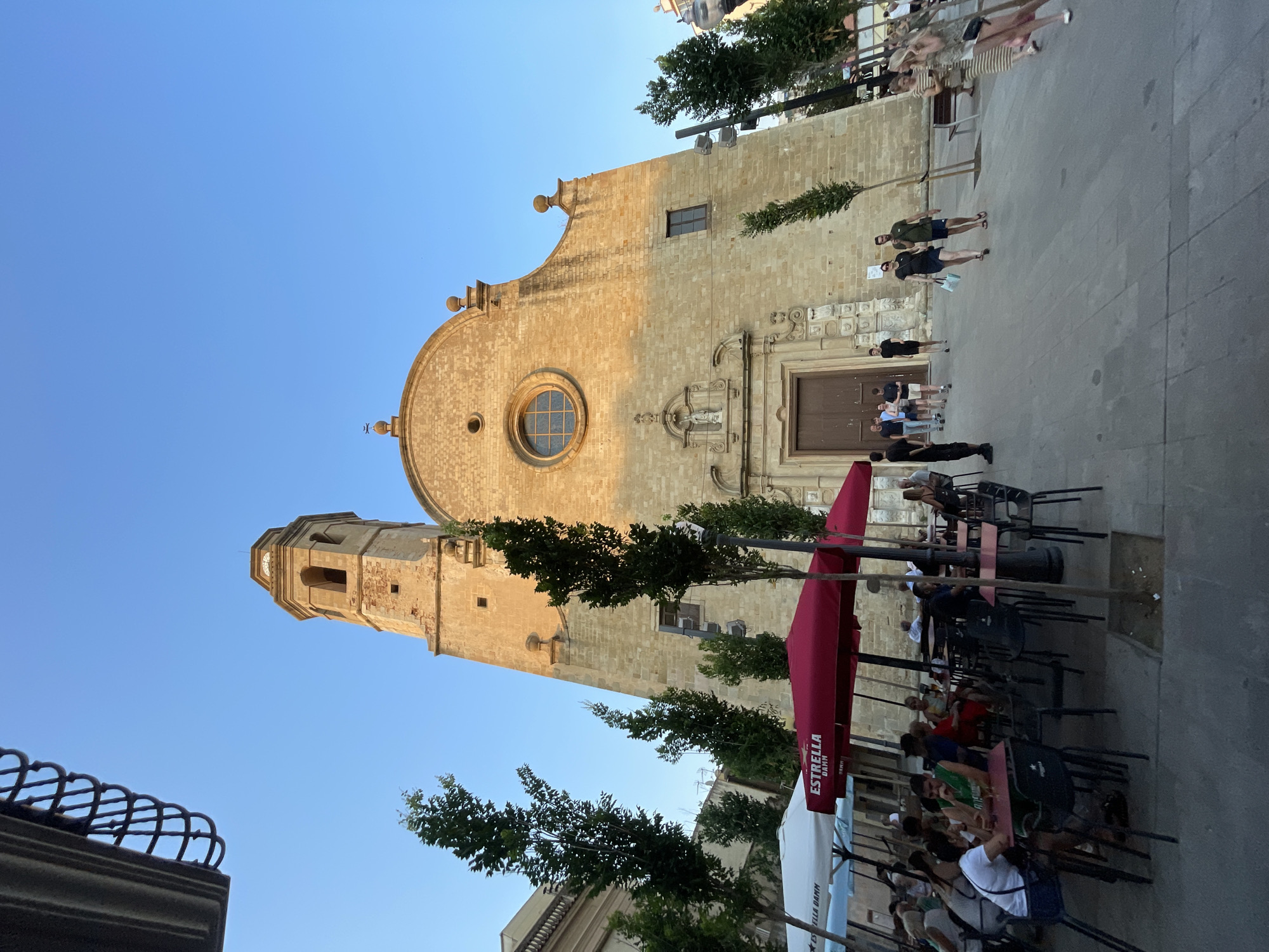 A wide shot captures the facade of a stone church with a bell tower in Calella, Spain. The church features a large wooden door, a circular window above it, and decorative stonework. In front of the church, a street-level plaza hosts an outdoor cafe with numerous tables and chairs covered by red and white umbrellas. Several people are seated at the tables, dining and socializing. Trees are planted along the sidewalk, and more people are walking in the plaza. The sky is clear blue, and the lighting suggests it is late afternoon or early evening, with the sun casting warm light on the church.