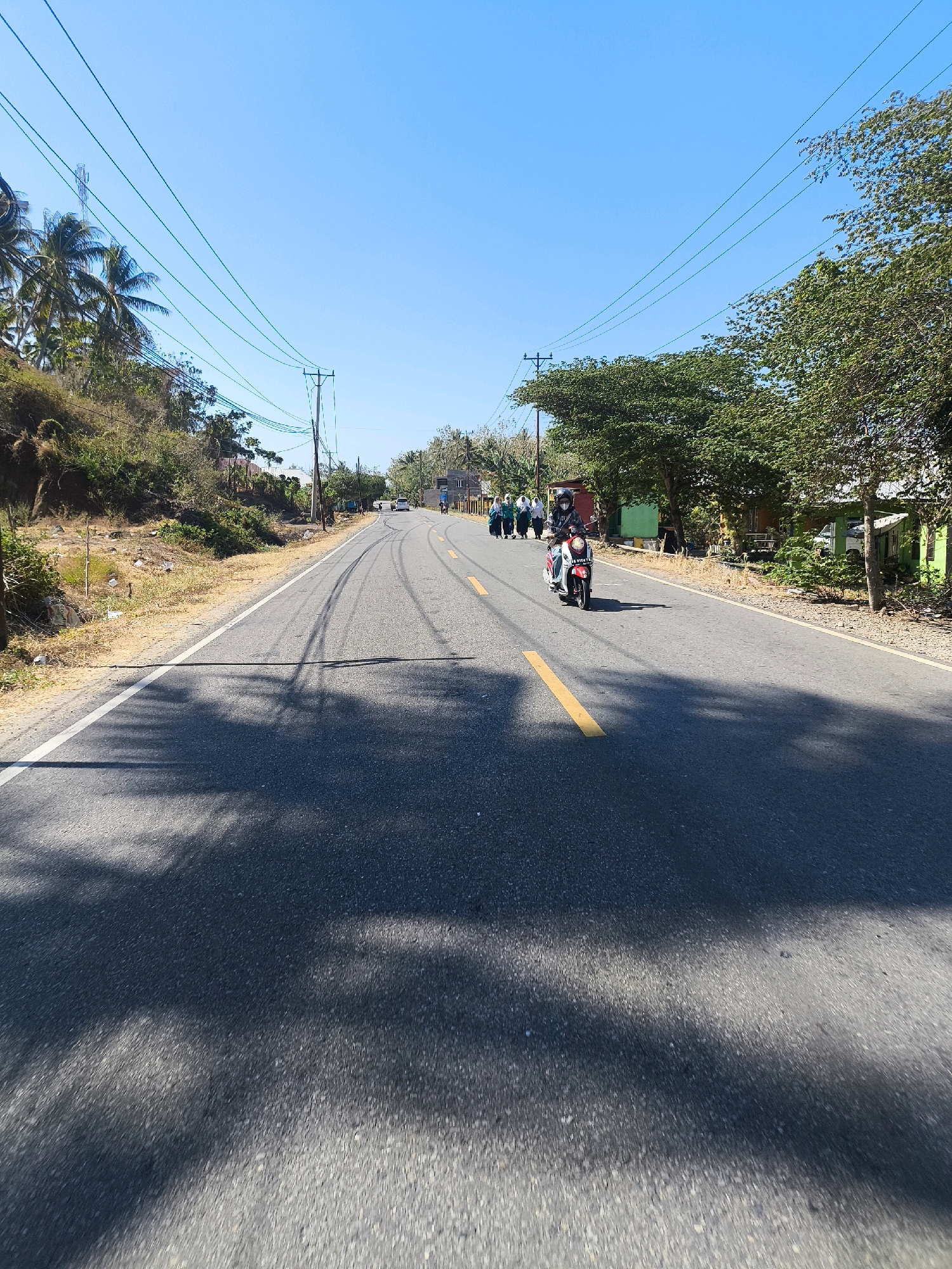 An eye-level, full shot captures a paved road in a rural area of Indonesia under a clear blue sky. The road is lined with vegetation and utility poles with power lines stretching across the frame. On the left side of the road, a grassy embankment is visible with some scattered debris. On the right side, a row of small buildings and trees can be seen.
In the middle ground, a scooter with a red and white body is being ridden by a person wearing a helmet. Following closely behind the scooter is a group of people, some of whom appear to be women wearing headscarves. In the distance, other vehicles are on the road, and there is a cell tower on the left side of the frame. The road has tire marks and a yellow dividing line. Shadows of trees are cast across the asphalt, indicating a sunny day. The overall scene depicts a typical day on a road in a semi-rural Indonesian setting.