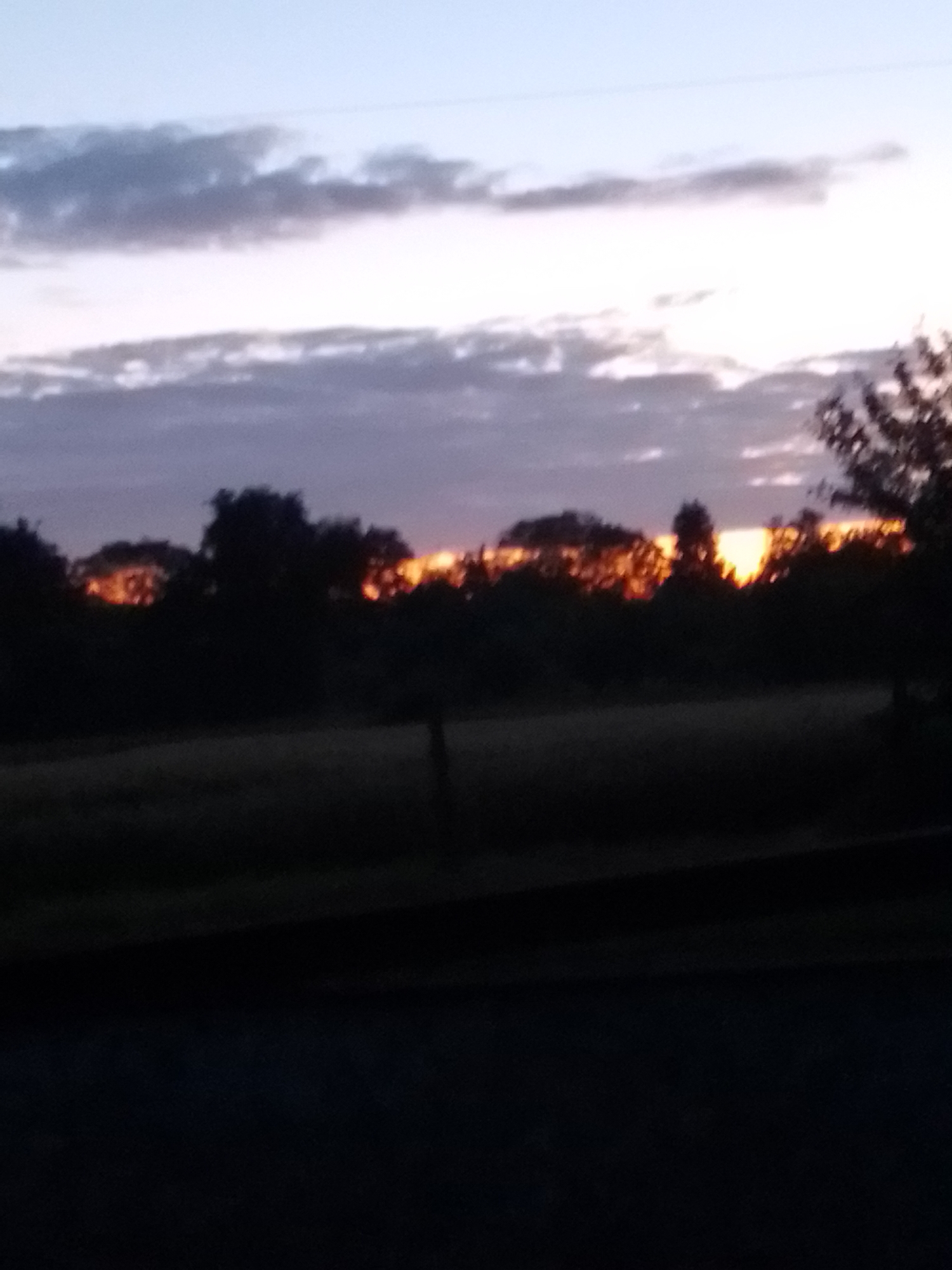 This is a photograph taken at dusk in Ustyvytsia, Ukraine. The image depicts a silhouetted landscape against a sky filled with scattered clouds. The horizon is lined with dark trees, with the sun's orange and yellow glow peeking through the gaps between them. The lower part of the image is very dark, suggesting it might be a foreground of asphalt or a dark surface. The overall impression is that of a rural or semi-rural setting as the day ends.