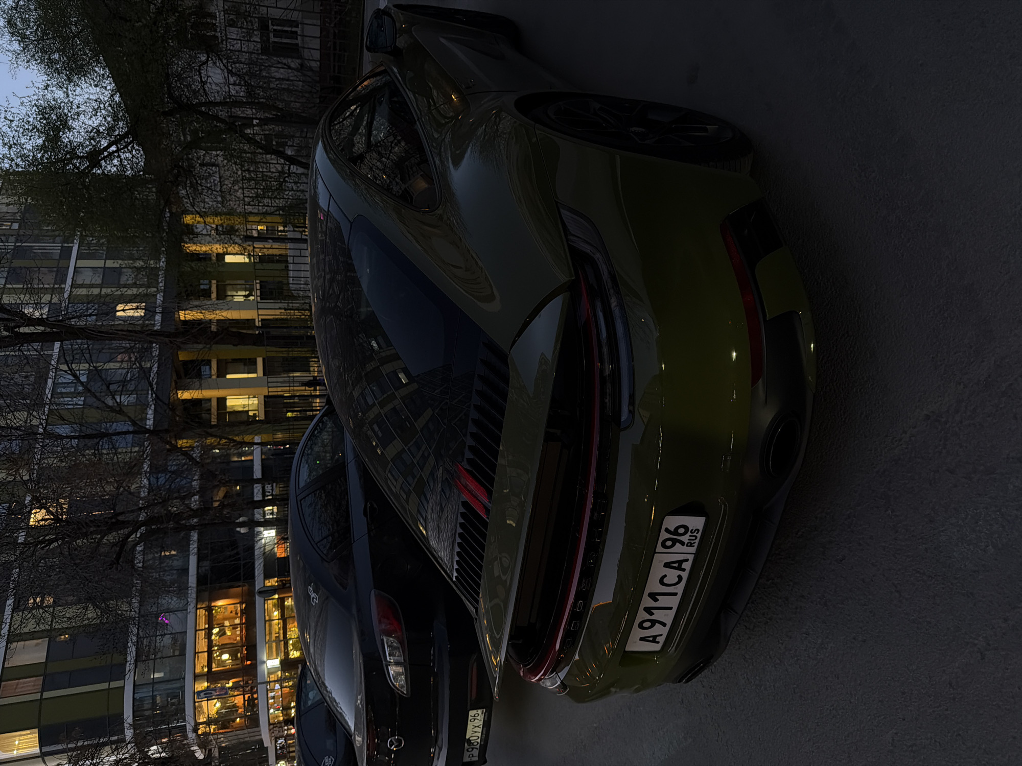 The vertical image, rotated 90 degrees clockwise, captures an urban street scene at dusk or early night in Yekaterinburg, Russia. The primary subjects are two parked cars: a distinct green Porsche 911 in the foreground and a dark-colored sedan (possibly a Mazda) partially visible behind it. No people are present.
The green Porsche is seen from its rear-right quarter, showcasing its sleek design, prominent taillight bar, and dual exhaust pipes. Its license plate, "A911CA 96 RUS," clearly identifies it as Russian, with "96" being the region code for Sverdlovsk Oblast, where Yekaterinburg is located. The adjacent dark sedan also displays a Russian license plate, "P980YX 96," confirming the same region.
In the background, bare-branched trees stand against a twilight sky, hinting at autumn or winter. Behind the trees, several modern, multi-story buildings line the street. Many windows of these buildings are illuminated with warm yellow and orange light, contrasting with the dark exterior and indicating activity within. The reflection of the buildings and lights is visible on the cars' surfaces, particularly the Porsche's sleek bodywork. The pavement or road surface is dark and appears slightly textured. The scene conveys a quiet, late-evening urban ambiance.