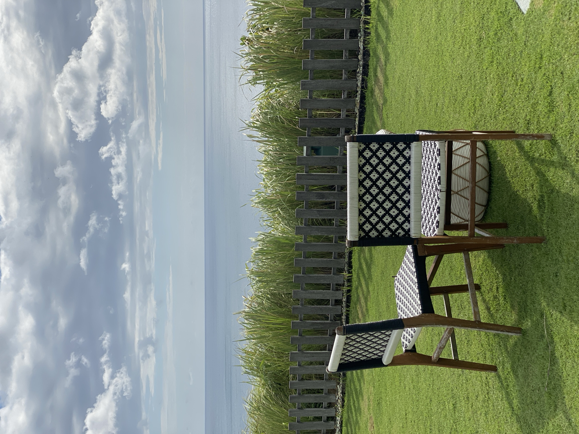 The image depicts an outdoor scene with two chairs on a manicured green lawn, facing a wooden fence that separates the lawn from a dense band of tall, grassy plants. Beyond the plants and fence lies a calm, blue ocean under a cloudy sky. The chairs are made of wood with woven seats and backs featuring a black and white geometric pattern. The left chair is angled slightly towards the viewer, while the right chair is more directly facing away. The wooden fence is composed of vertical, weathered planks. The overall setting suggests a serene, coastal location, likely a resort or a scenic viewpoint. The location is identified as being in Kuta Selatan, Indonesia.