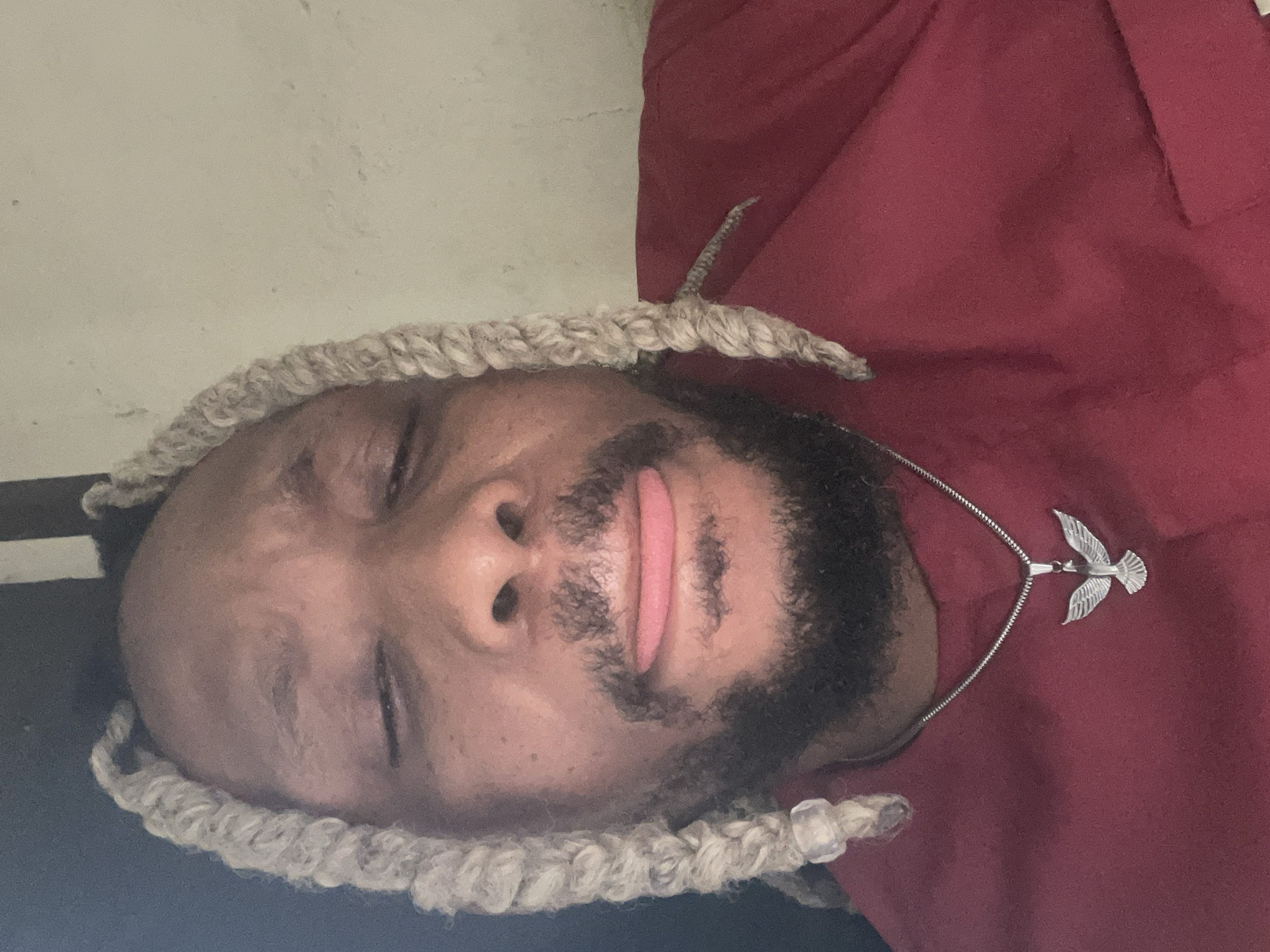 A photograph depicts a man from the chest up wearing a maroon top. A silver-colored chain with a bird pendant is around his neck. He has a beard. He wears beige/grayish dreadlocks. His eyes are closed, and his tongue sticks slightly out between his lips. The photograph was taken indoors, with one wall a light beige and the other a dark color. The location cannot be verified without additional information.
