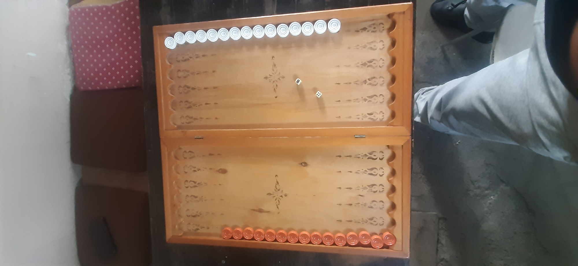 A top-down, slightly angled view captures a wooden backgammon board with its lid open, revealing a game in progress. The board is positioned on a dark, textured surface, possibly concrete or stone. Orange checkers are stacked along the left edge of the board, while white checkers are lined up on the right edge. Two white dice are visible on the board, one showing a '3' and the other a '6'. The environment surrounding the board is dim, with some indistinct objects in the background, including what appear to be cushions on a surface. The lower portion of the image shows a person's legs clad in light grey pants, with dark shoes visible on the right. The overall scene suggests a casual setting, possibly a home or a recreational area. The location of Yessentukskaya, Russia, is not discernible from the visual information in the image.