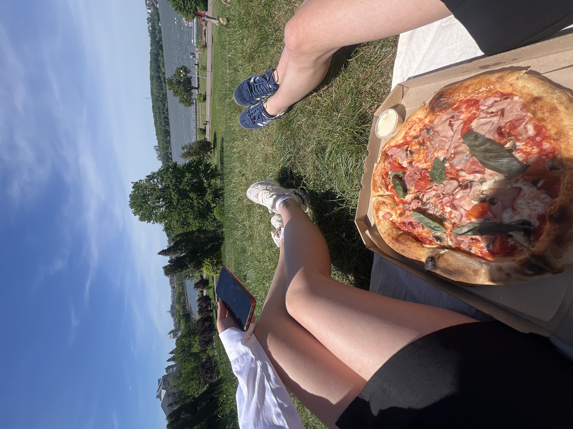 On a sunny day in Ternopil, Ukraine, two people are sitting on the grass, enjoying a pizza picnic by the lake. One person is wearing blue sneakers and black shorts. Next to them is a pizza in a cardboard box with a small container of dipping sauce. The pizza has ham, tomatoes, and basil. The other person is holding a cell phone and wearing white sneakers, black shorts, and a white shirt. The lake is visible in the background with trees along the shore and a clear blue sky above.