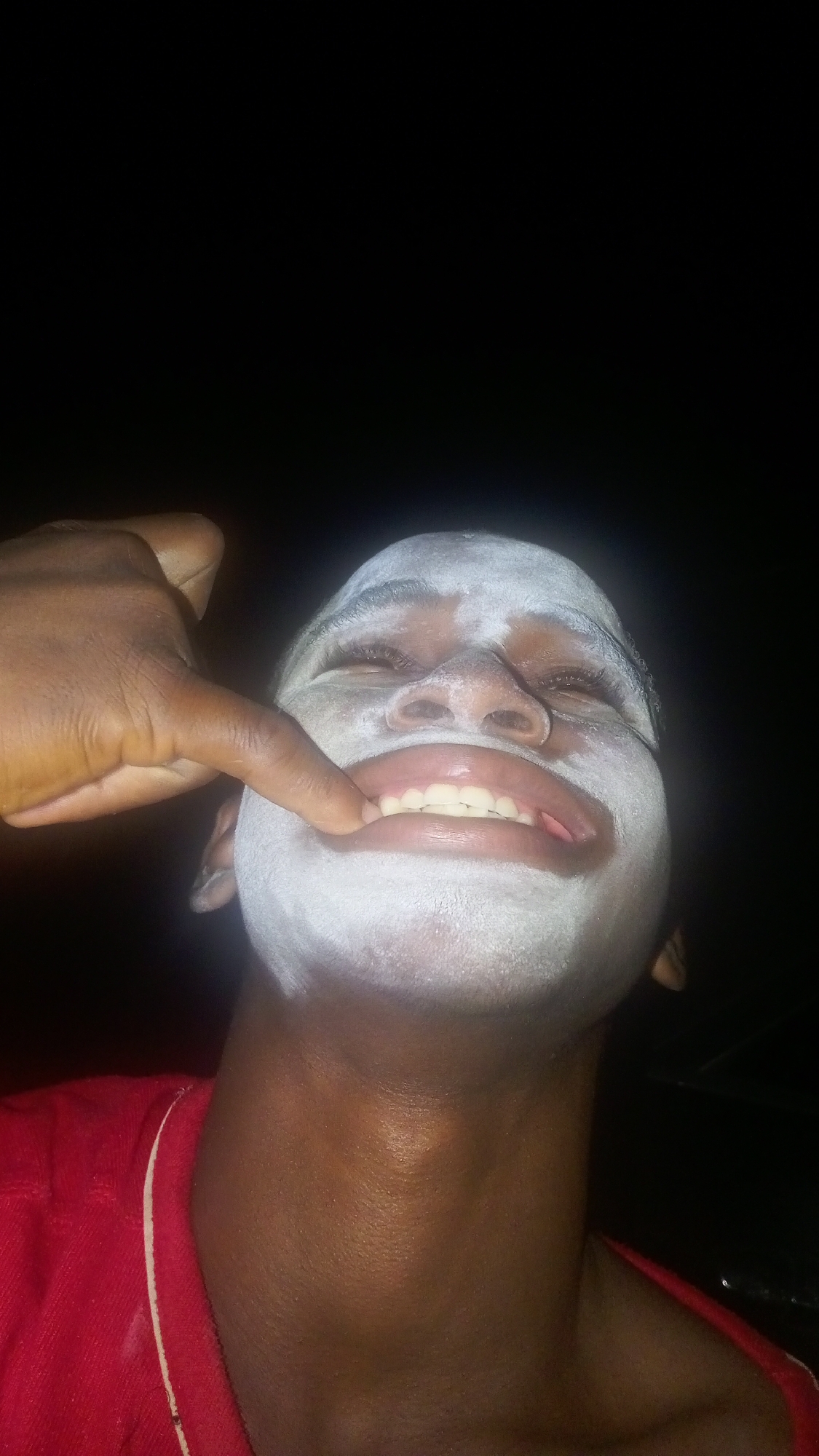 A low-angle photograph depicts a person with dark skin, presumably male, with white cream or paint applied to their face, extending to the forehead and chin. The person is wearing a red shirt with a white trim at the collar. One finger touches the lower lip. The background is black. The location is inferred to be Onitsha, Nigeria.