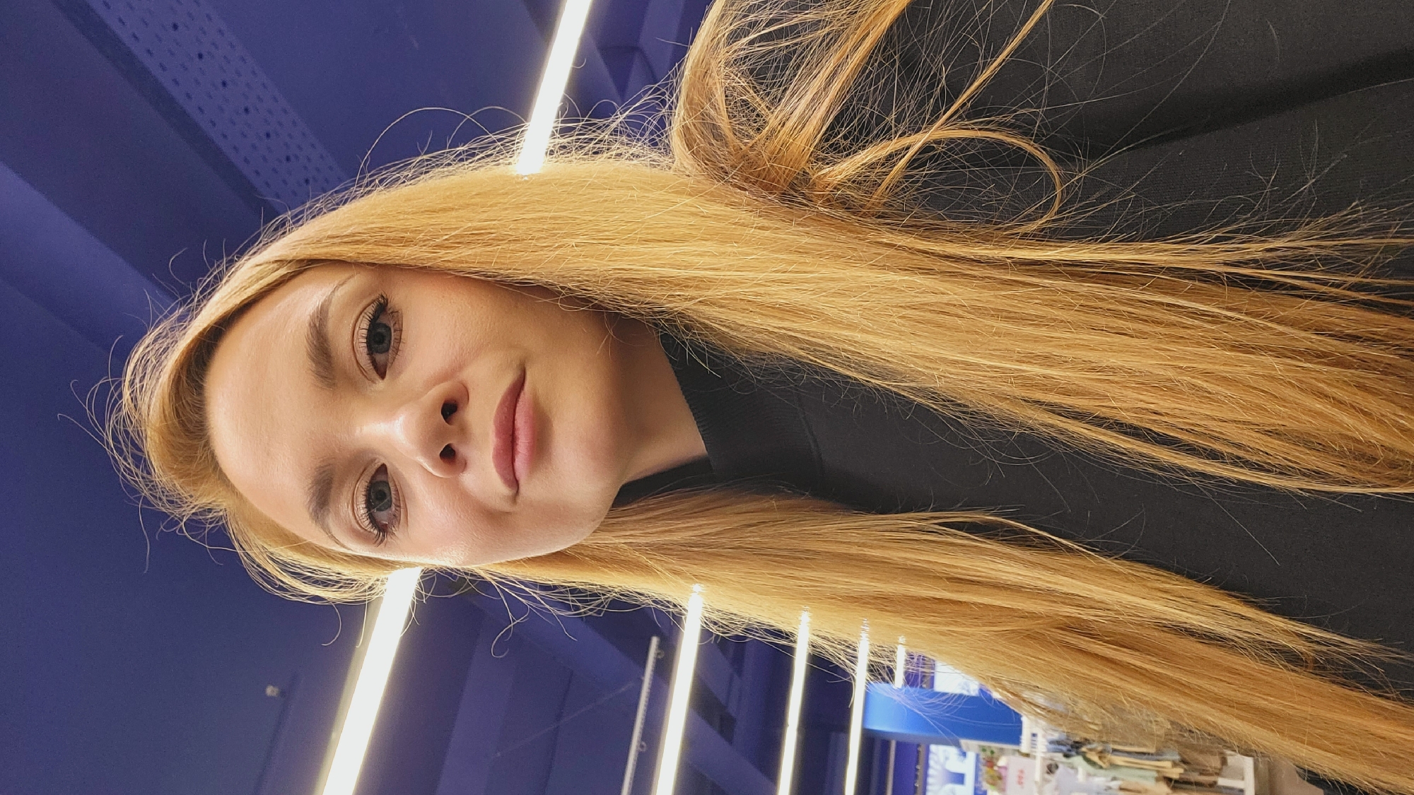 A young woman with long, straight, light blonde hair is captured in a low-angle selfie. Her head is tilted slightly, and she looks directly into the camera with a neutral to slightly pleasant expression. Her eyes are a light blue or blue-grey, reflecting the bright artificial lights in the background. She has fair skin and naturally colored lips, which are slightly parted. She is wearing a dark, possibly black, high-necked top.
The background features a deep blue ceiling or wall, indicating an indoor setting. Multiple bright, linear fluorescent light fixtures are visible, running parallel across the upper portion of the frame. These lights illuminate the scene and create distinct reflections in her eyes. The overall environment suggests a modern commercial or public space, possibly a store or exhibition hall, as hinted by blurry elements and shelving visible in the far distance on the right side of the main image. The image was taken in Chaikovsky, Russia. No discernible text is visible in the frame.