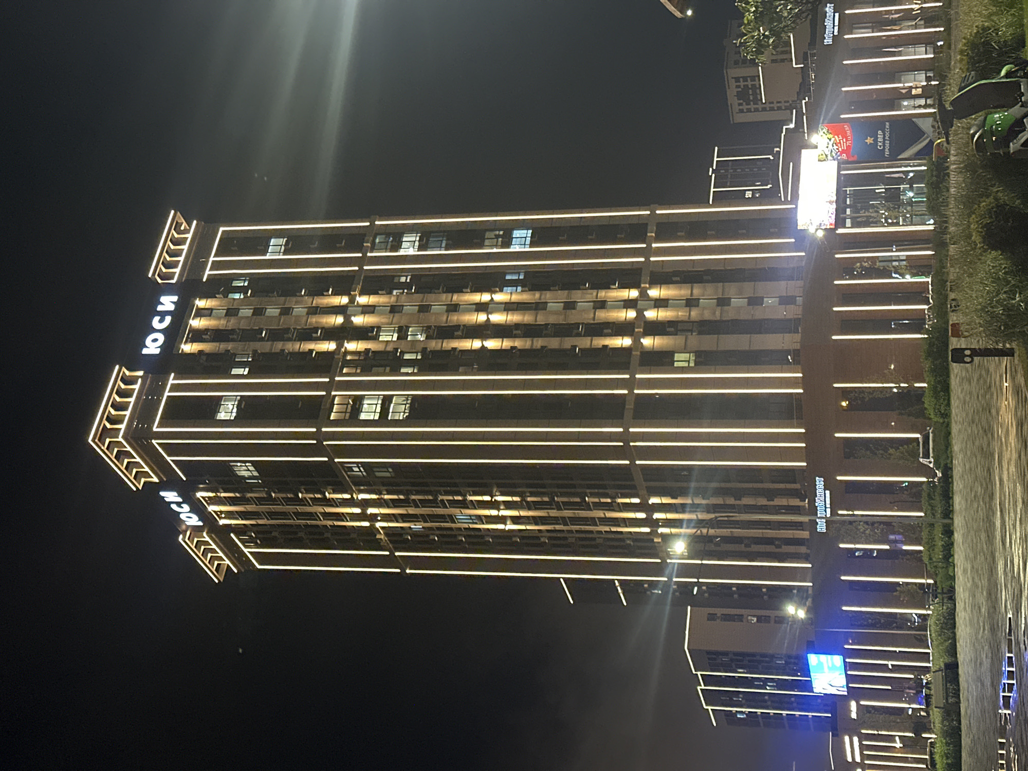 The image captures a nighttime scene of a tall building in the city of Stavropol, Russia. The building, adorned with rows of horizontal lights, dominates the frame and displays the name "ЮСИ" vertically on its facade. Adjacent to the building is a park-like area with trees and manicured landscaping. Further buildings, also illuminated, are visible in the background. A screen or billboard displays "СКВЕР ГЕРОЕВ РОССИИ," suggesting a memorial square dedicated to heroes of Russia. The overall setting suggests an urban environment with modern architecture and illuminated nightscapes.