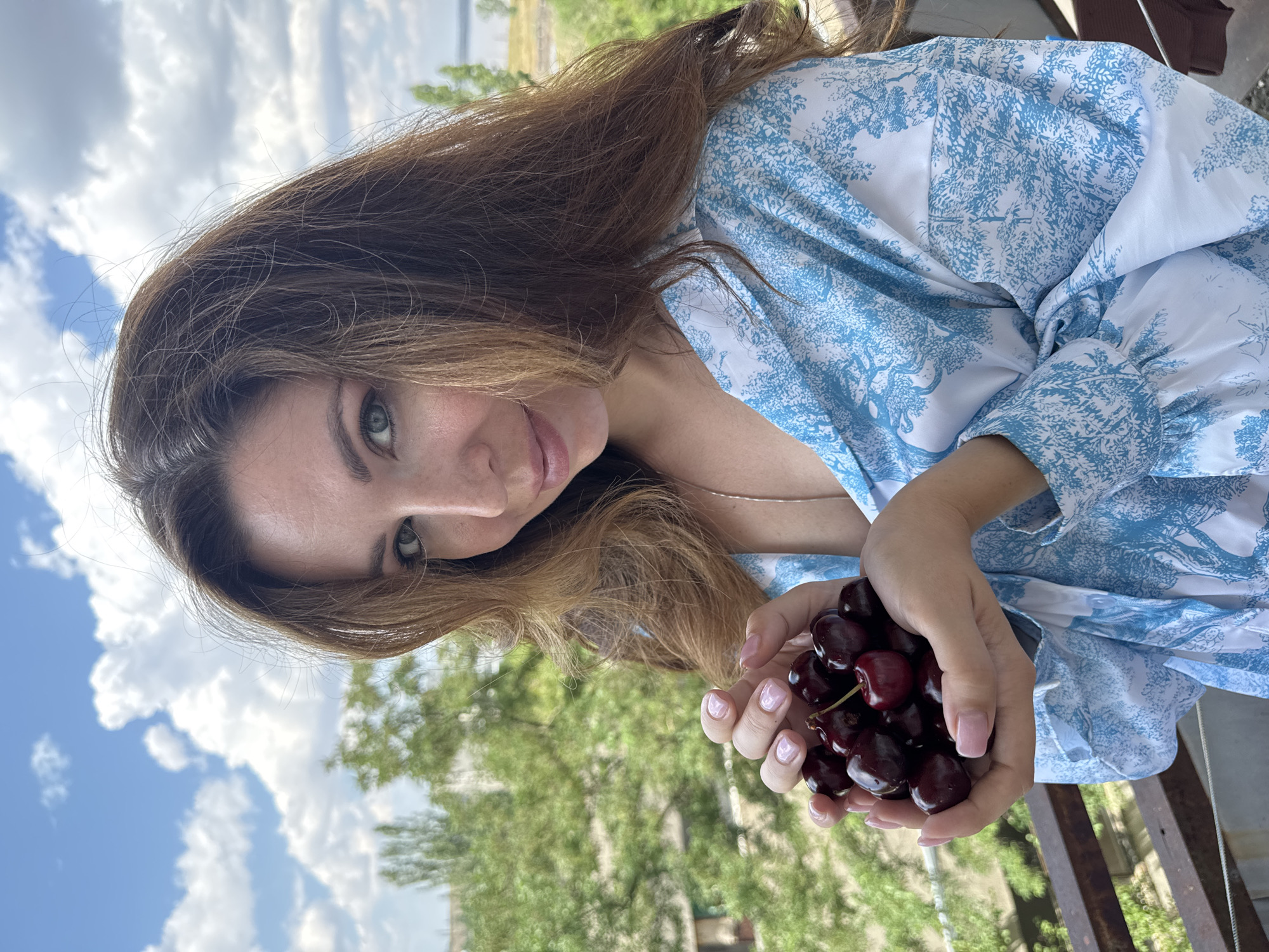 A woman with long brown hair stands on a balcony, holding a handful of dark red cherries. She is wearing a blue and white patterned shirt. The background shows trees and a cloudy sky. The scene appears to be in Odesa, Ukraine, based on the provided information.