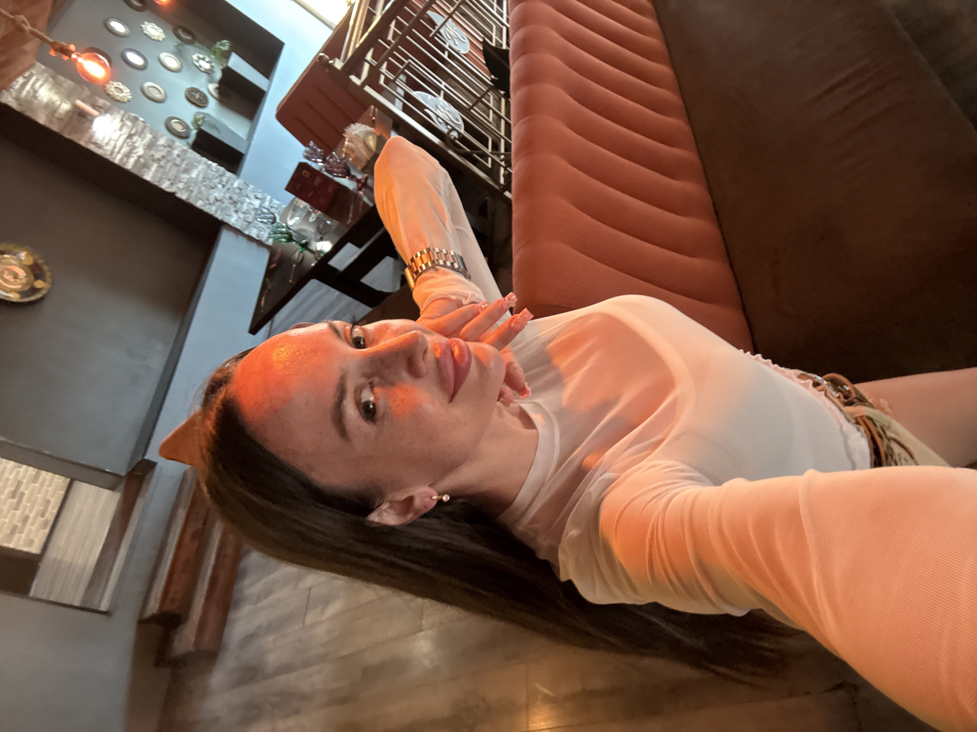 An outdoor shot shows a woman in a white sheer top sitting on a velvet couch. Her left hand is resting on her chin while her right hand is extended outwards. She has long dark hair and fair skin with freckles. The background features a restaurant interior with wooden floors, brick walls, and ambient lighting from decorative lamps. In the distance, a table with glasses and chairs is visible. There is no visible indication of the image being taken in Saint Petersburg, Russia.