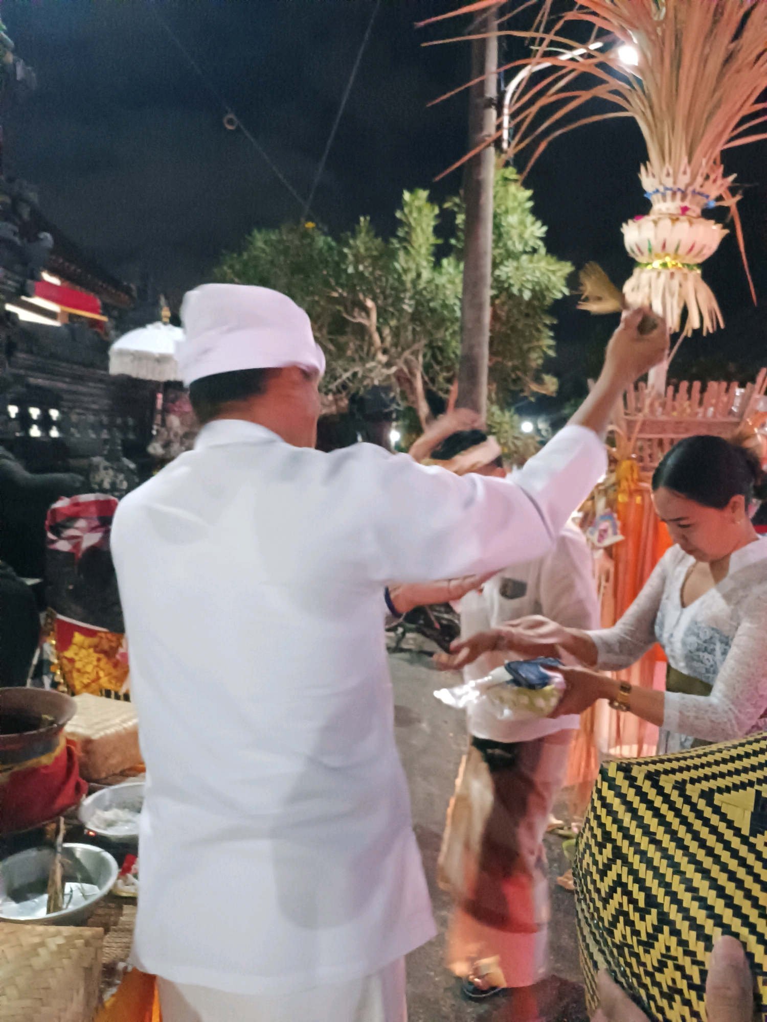 The image depicts an outdoor evening ceremony, likely at a temple in Kediri, Indonesia, given the traditional attire and offerings. In the foreground, a person wearing a white garment and a white head covering is partially visible from the back, with their right arm raised. To the right, a woman in a white, lace-trimmed traditional outfit, with her hair tied back, is receiving an offering. She is wearing a gold bracelet. The offering appears to be in a clear plastic container. Behind them, other individuals in similar white attire are present, one of whom is partially obscured. The environment includes trees, a structure resembling a temple gate with red and black detailing on the left, and a large, decorative palm frond arrangement on a pole to the right, illuminated by a light source. Various offerings and baskets are visible on the ground in the lower left and right of the frame, including woven baskets and bowls containing what appear to be food or other ritual items. The ground surface is dark and appears to be paved.